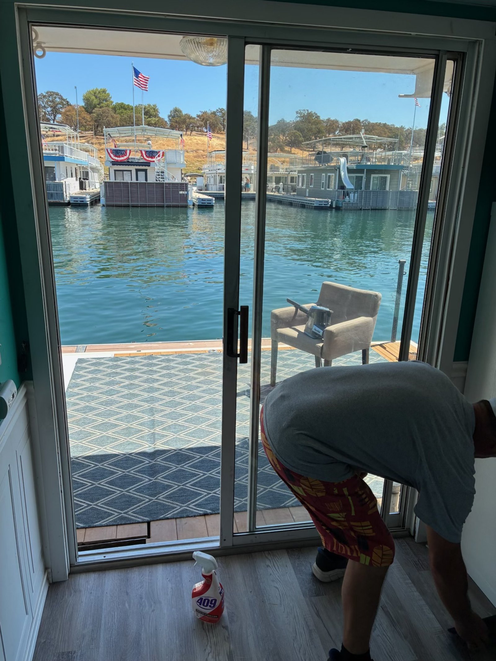 View through a glass sliding door showing a dock with houseboats decorated with American flags, and a man inside bending over near the door with a spray bottle on the floor.