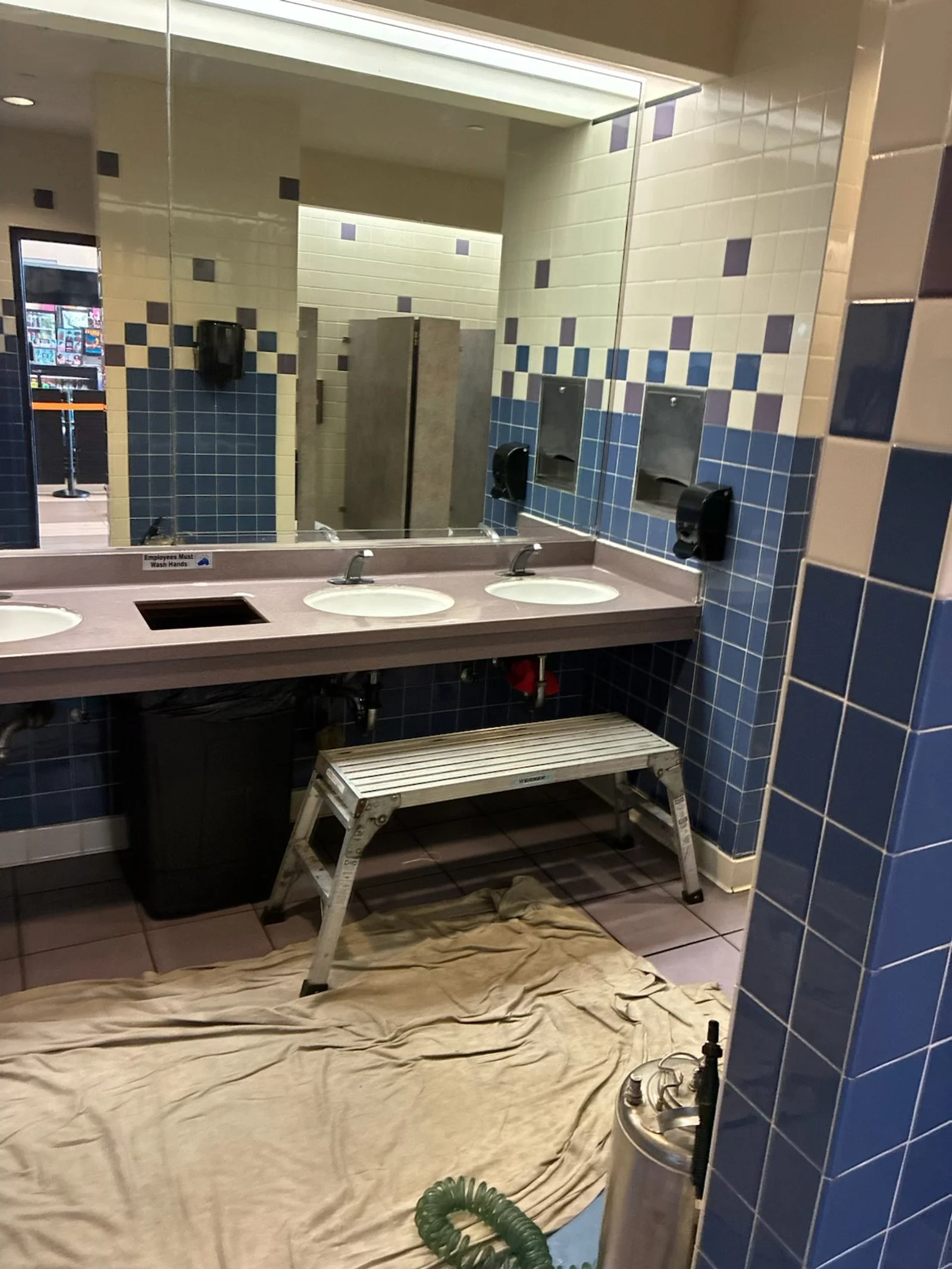 Public restroom with a triple sink, large mirror, and blue tiled walls. A step ladder and a sheet are on the floor, and a fire extinguisher is nearby.