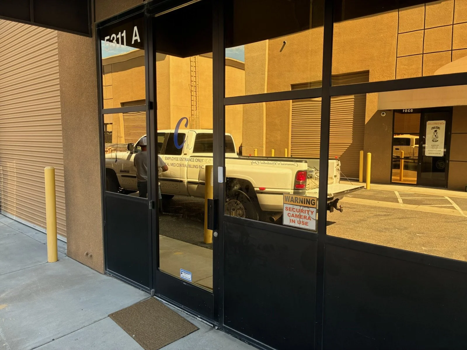 Reflection of a white pickup truck in a glass door with a security warning sign, yellow bollards outside a commercial building.