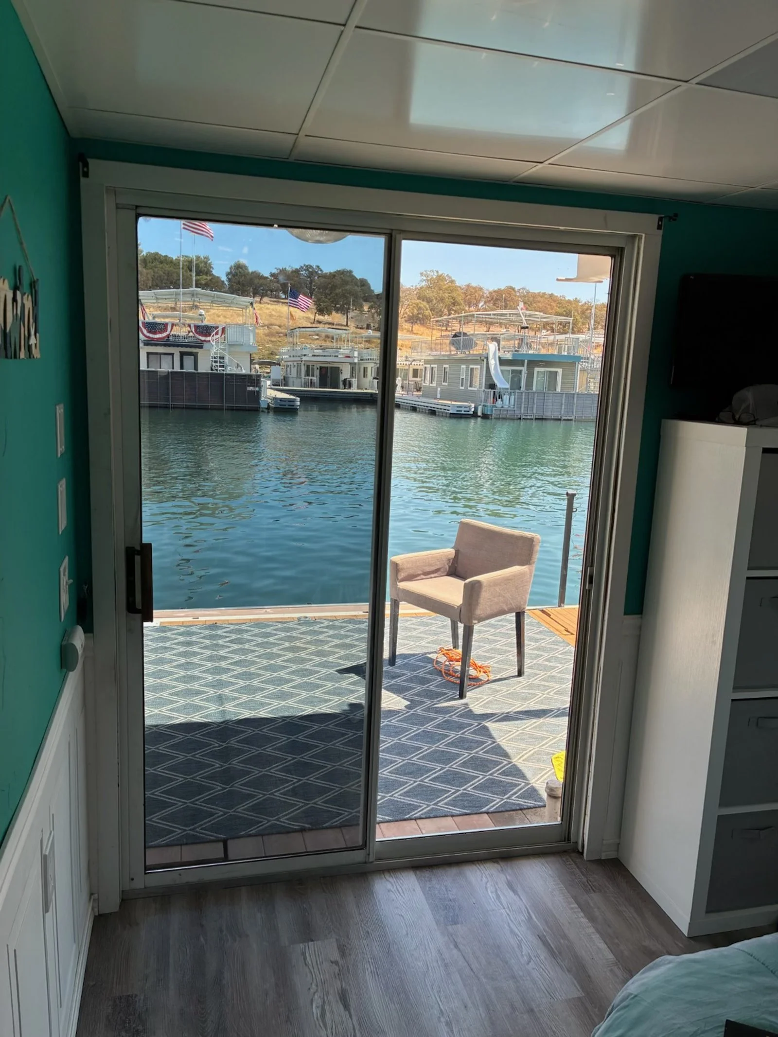 View of houseboats and water from inside a room with sliding glass door, outdoor chair on a patterned outdoor rug on dock, water, and houses with American flags in the background.