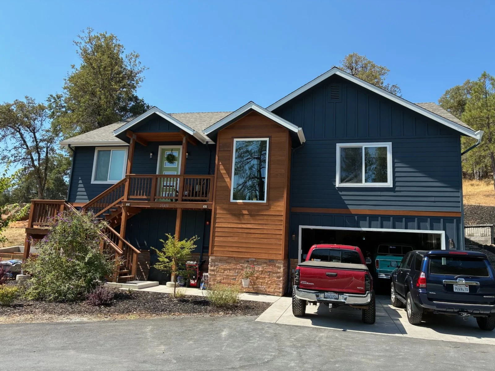 A modern two-story house with blue siding, wood accents, and a multi-angled roof. It has a small front porch with stairs, and a driveway with several parked vehicles, including a red truck, a black SUV, and a teal van. There are plants and trees arou