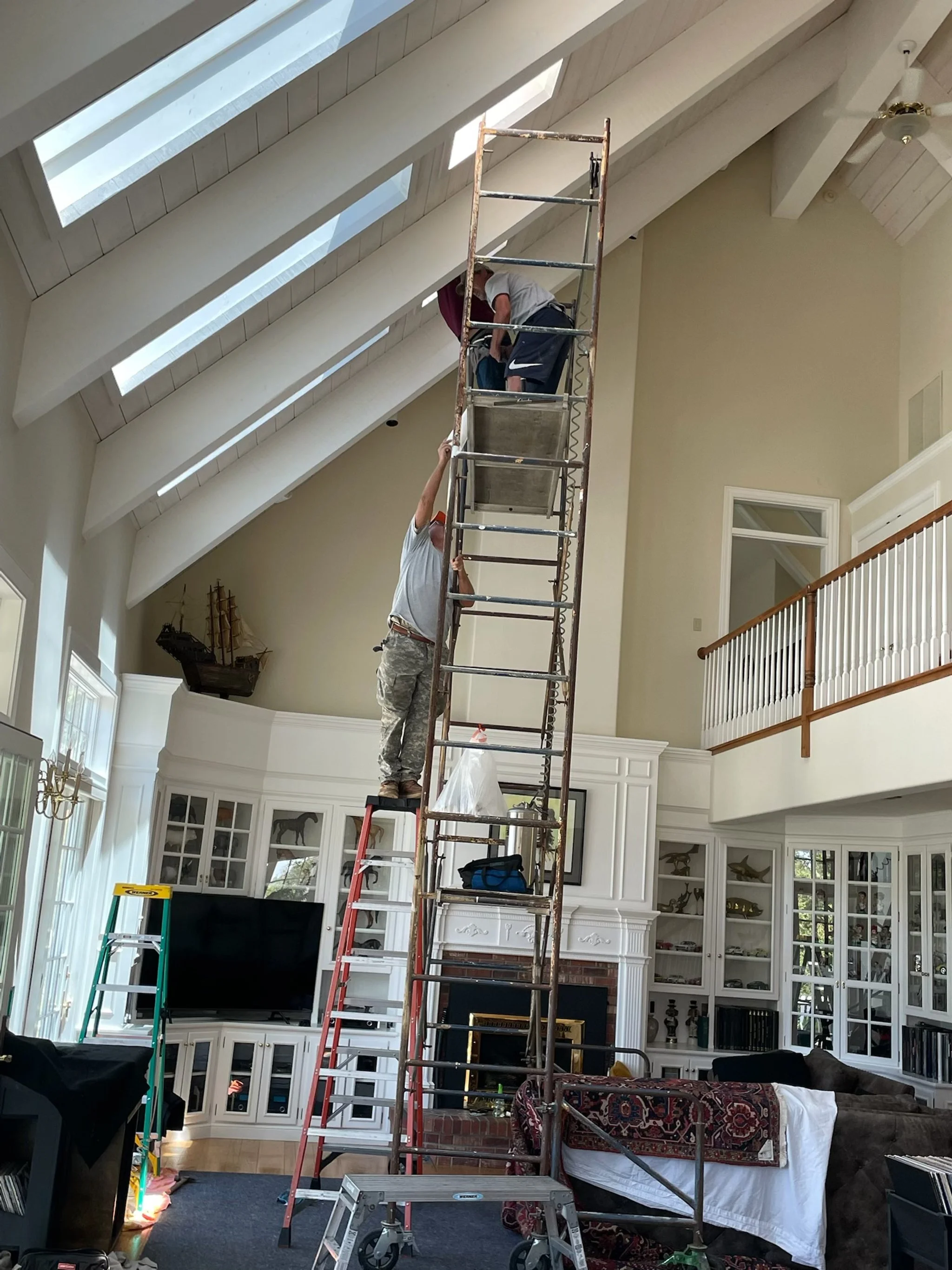 Two workers installing or repairing something in a room with a vaulted ceiling and skylights. They are standing on scaffolding, one on the ground and one on the upper platform.
