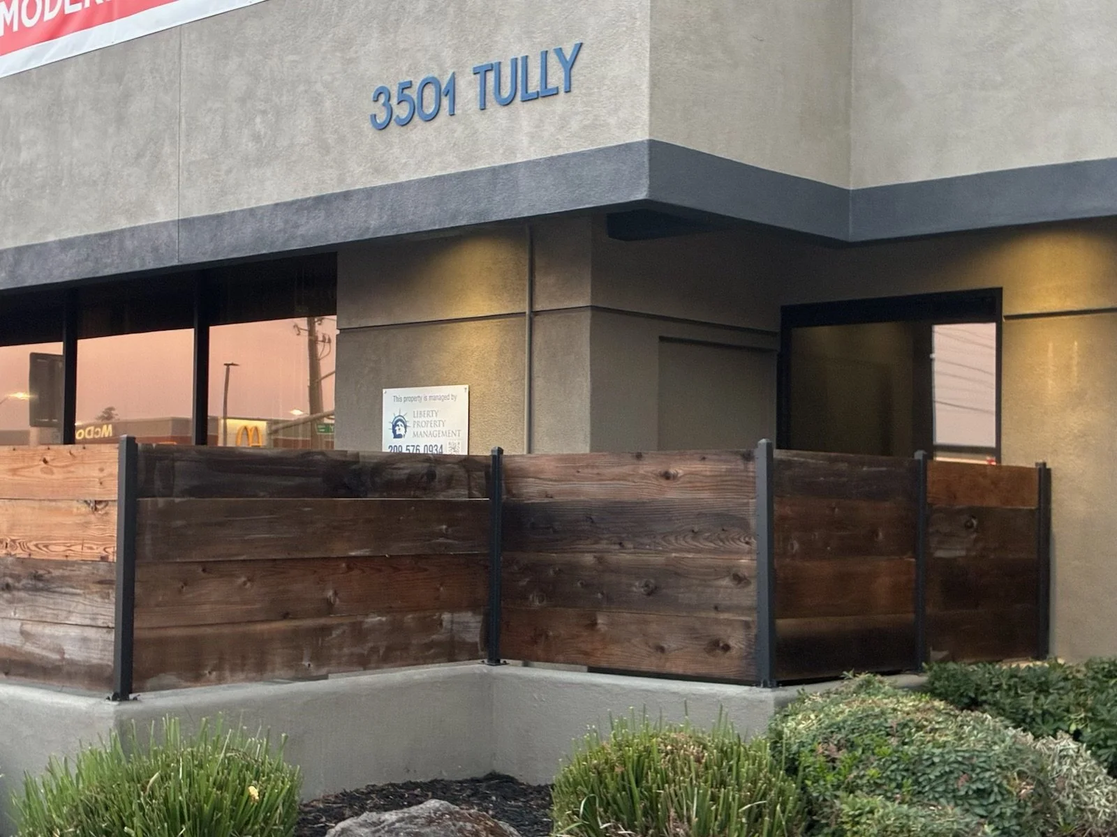 Corner of a commercial building with the address 3501 Tully in blue letters, a fenced outdoor area with wooden panels, and windows reflecting a nearby McDonald's and other buildings.