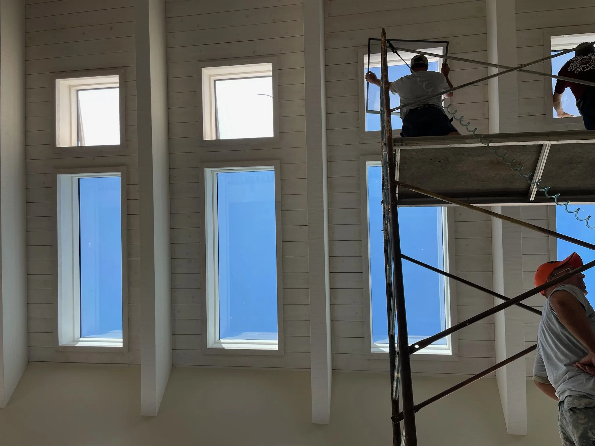 Construction workers on scaffolding installing or working on interior windows of a building with a high ceiling and multiple rectangular skylights.