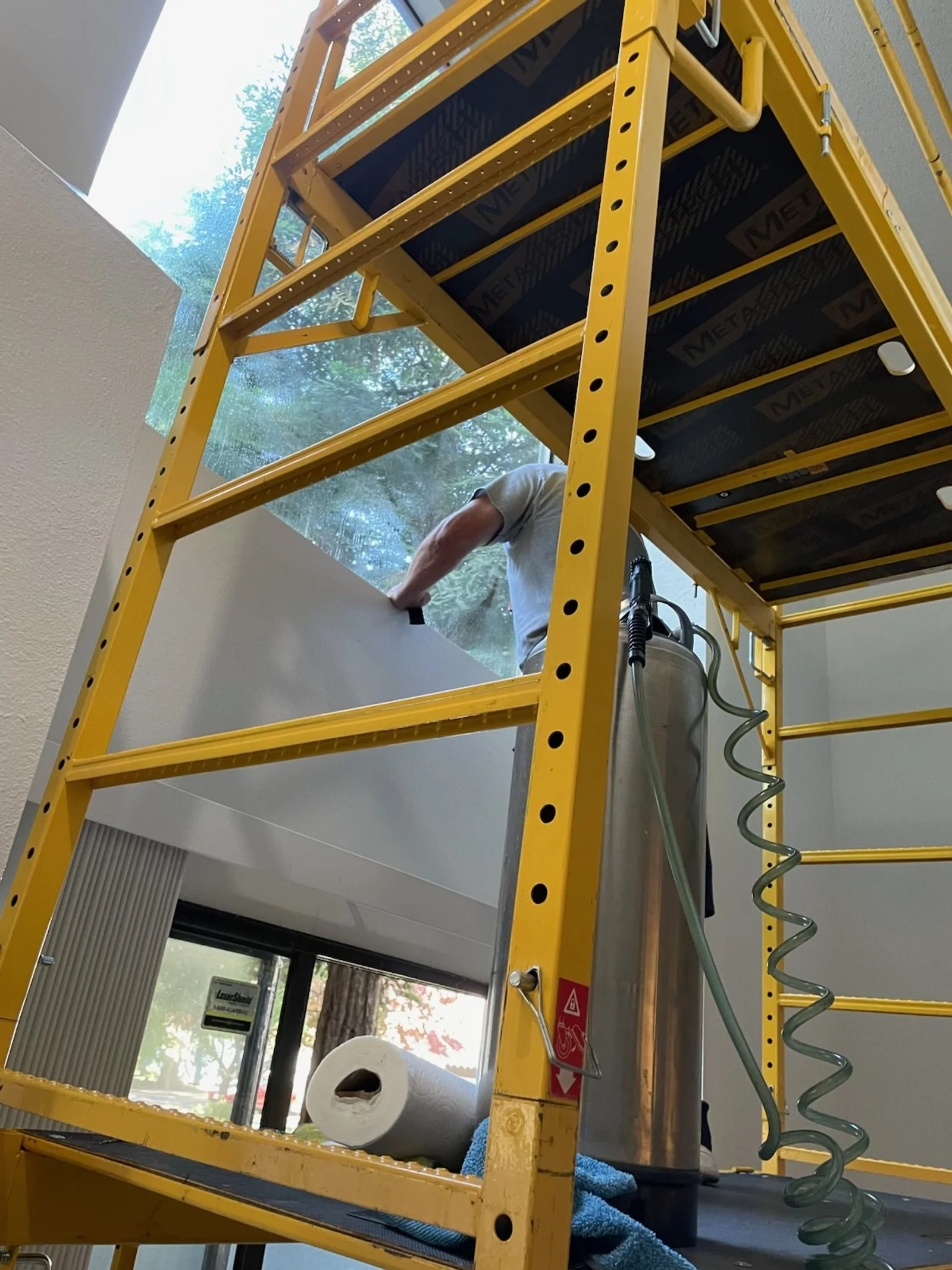 A person working on the inside of a yellow stepladder near a wall with a window, with cleaning supplies like a paper towel roll visible.