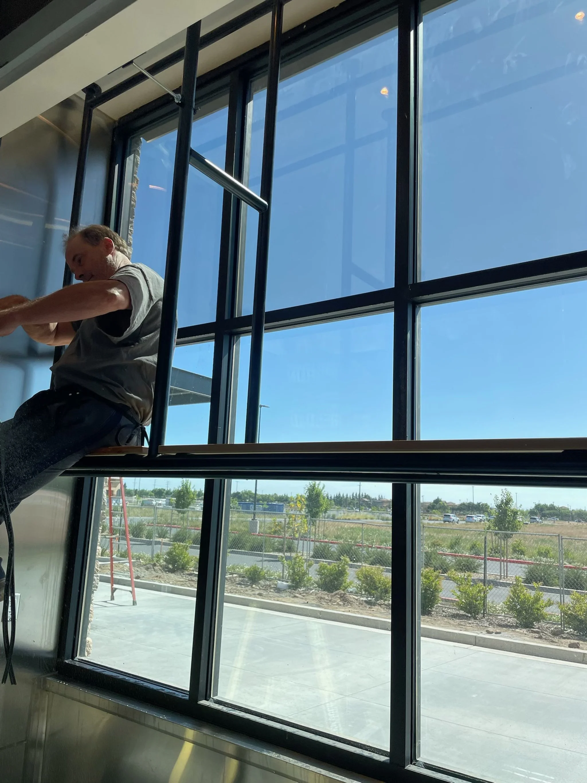A person working on a large window inside a building with a view of a parking lot, fence, and trees outside on a sunny day.