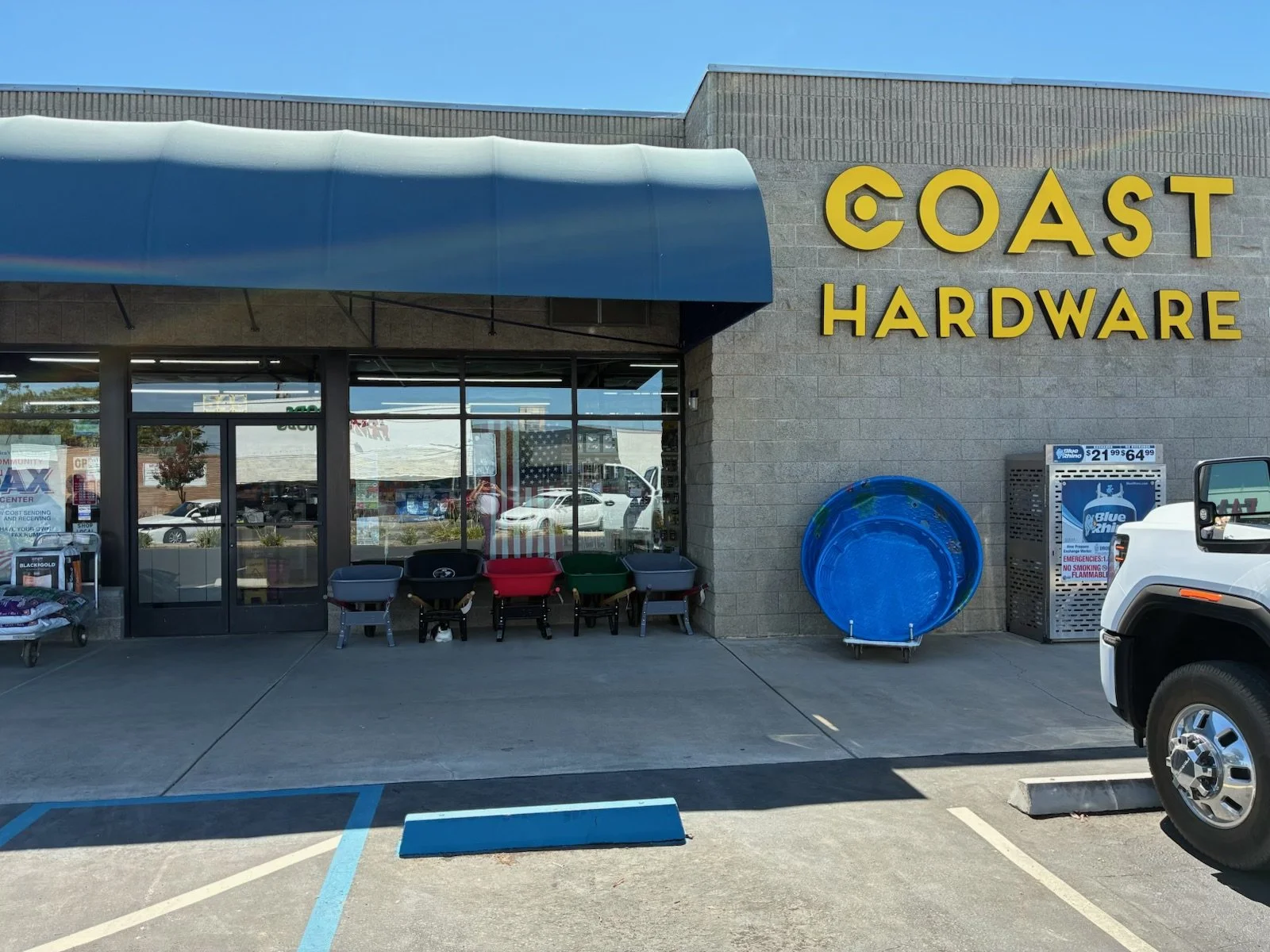 Exterior of Coast Hardware store with yellow sign, outdoor chairs, a large blue round item, and a vending machine.