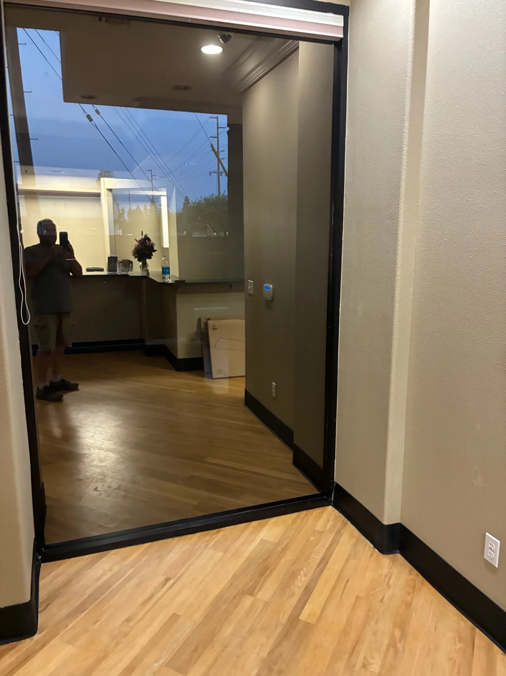 Interior view of a room at dusk with hardwood floors and a glass door leading outside, a person taking a photo using a smartphone, a reception desk with a flower arrangement and water bottle, and a person reflected in the glass door.