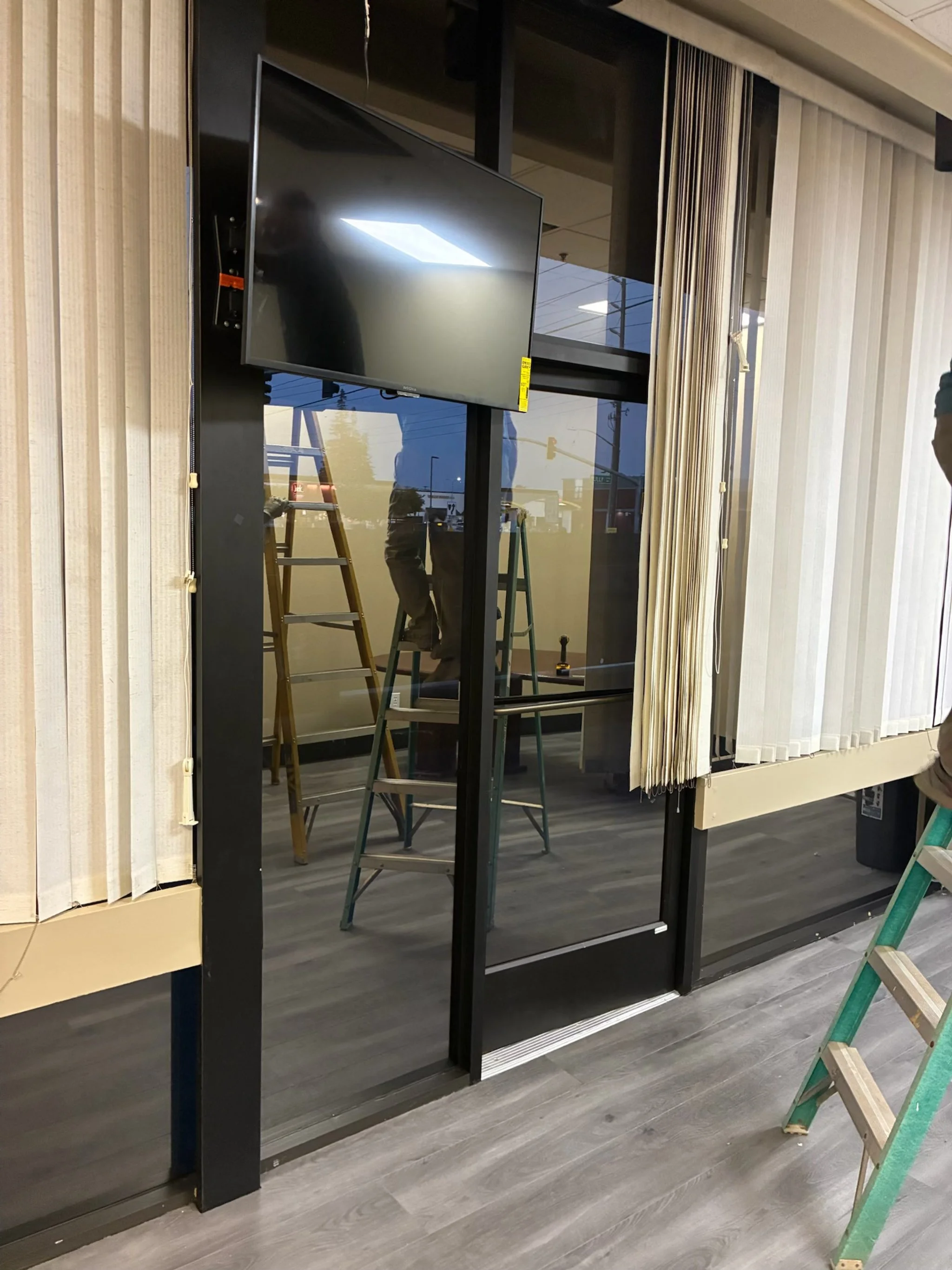 Interior of a commercial space under renovation with a glass door, vertical blinds, a wall-mounted TV, and ladders reflected in the glass.