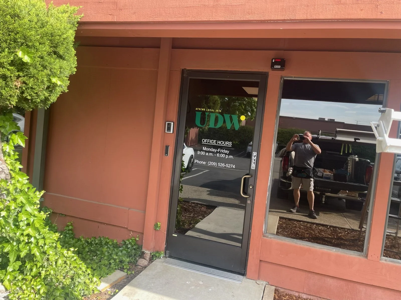 Photo of a storefront with a glass door and window, reflecting a person taking a picture, and on the door are the business hours and contact information. The building is painted a reddish-brown color and has greenery on the left side.