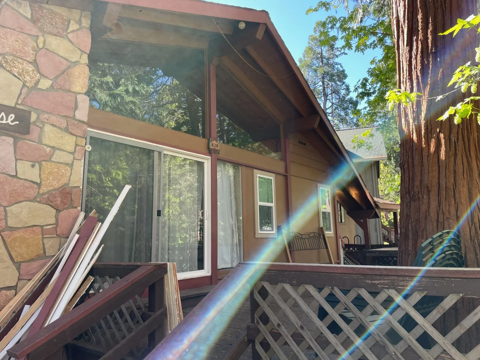 View of a house with large windows and a stone wall on the left, surrounded by trees, with sunlight and lens flare effects.