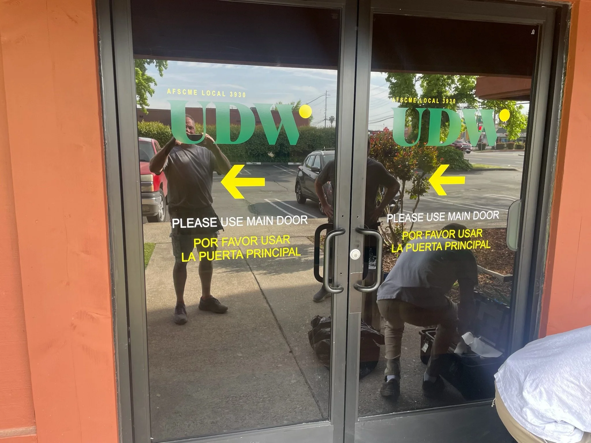 Double glass doors with yellow and white signage indicating to use the main door, with reflections of two men and a parking lot outside.