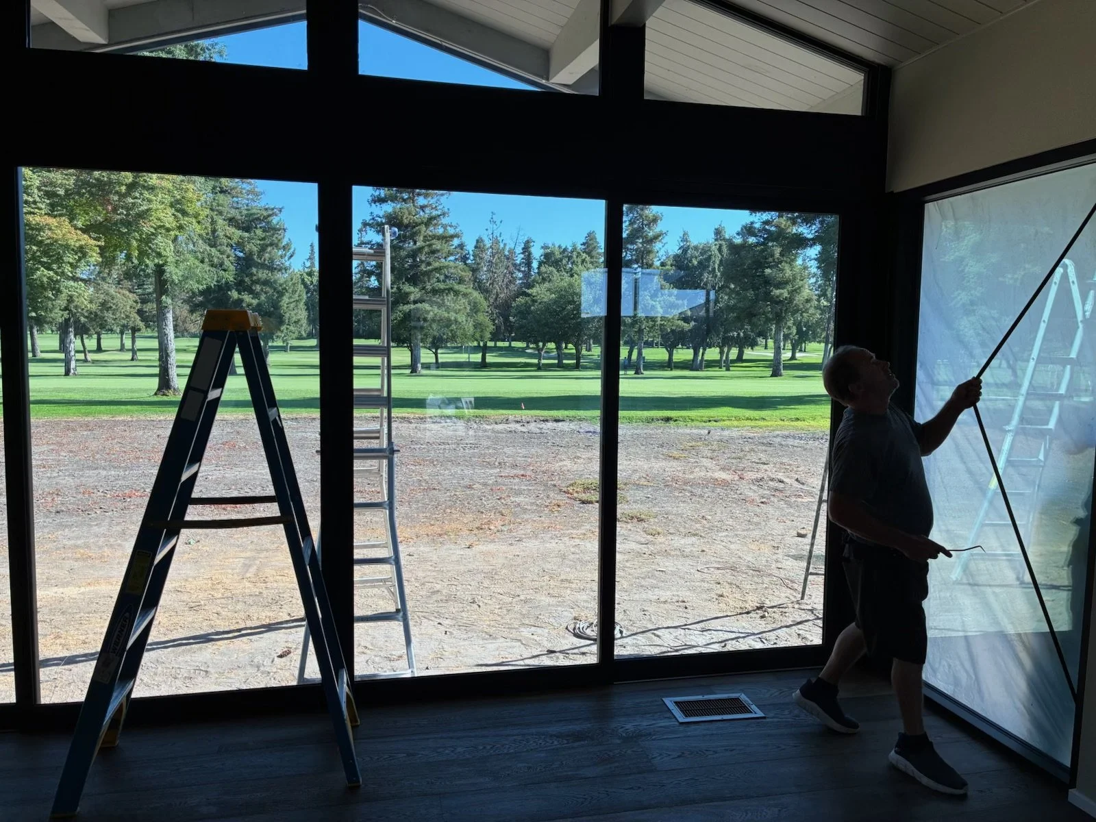 Person cleaning large glass window or door inside a building, with ladders outside visible through the window, overlooking a field with trees.