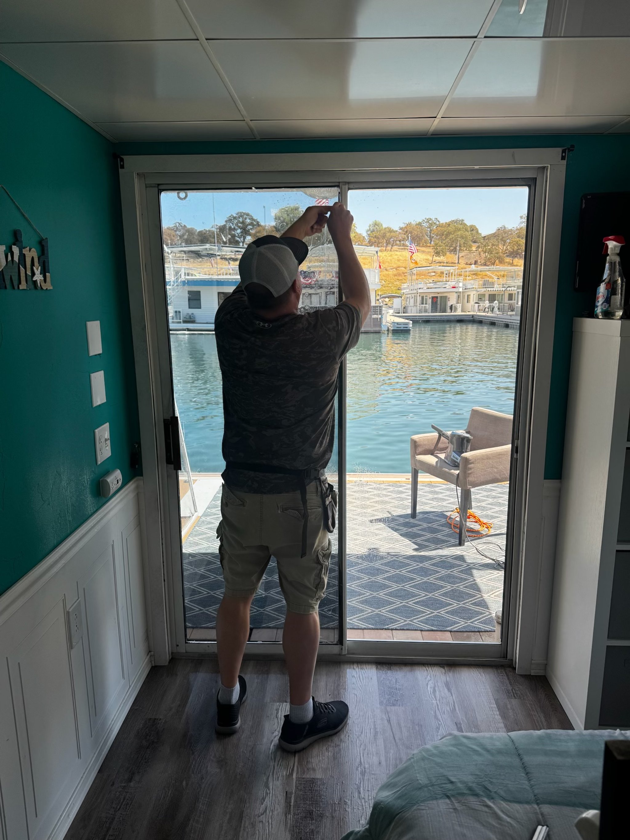 A man standing inside by a sliding glass door, hanging an object outside on a dock. Meadow and trees are visible across the water. The interior has teal walls and wood flooring.