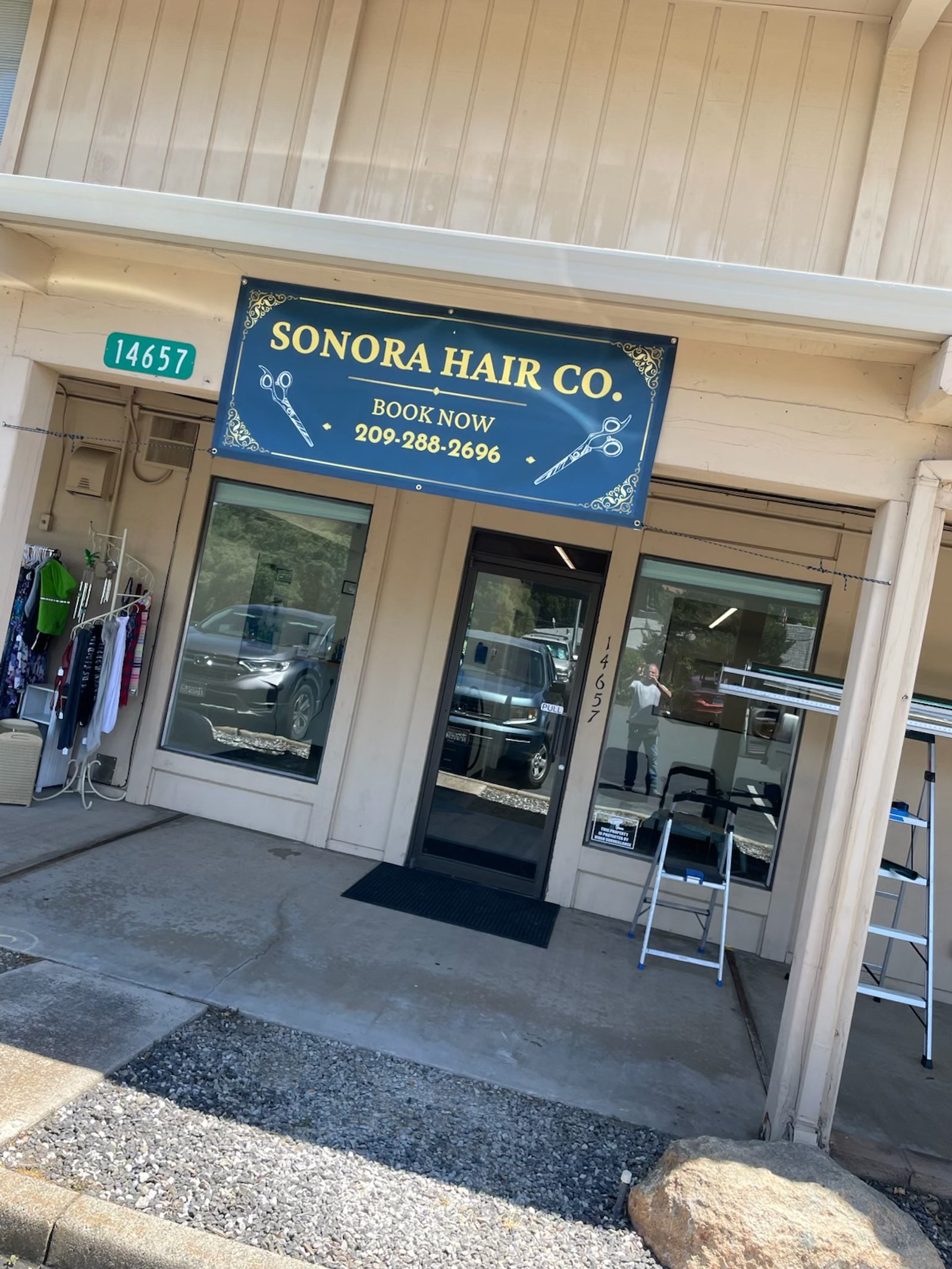 Front of a hair salon called Sonora Hair Co. with a blue sign, large windows, and an entrance door. There is a small display of clothing and a ladder outside.