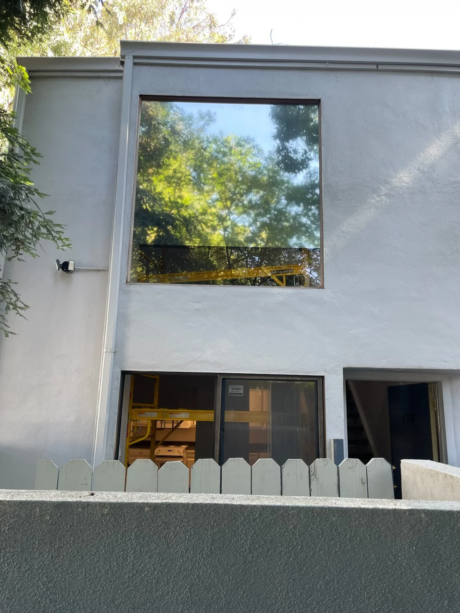 A two-story building with large glass windows, construction equipment visible inside, and a white picket fence in front. Trees reflect in the upper window.