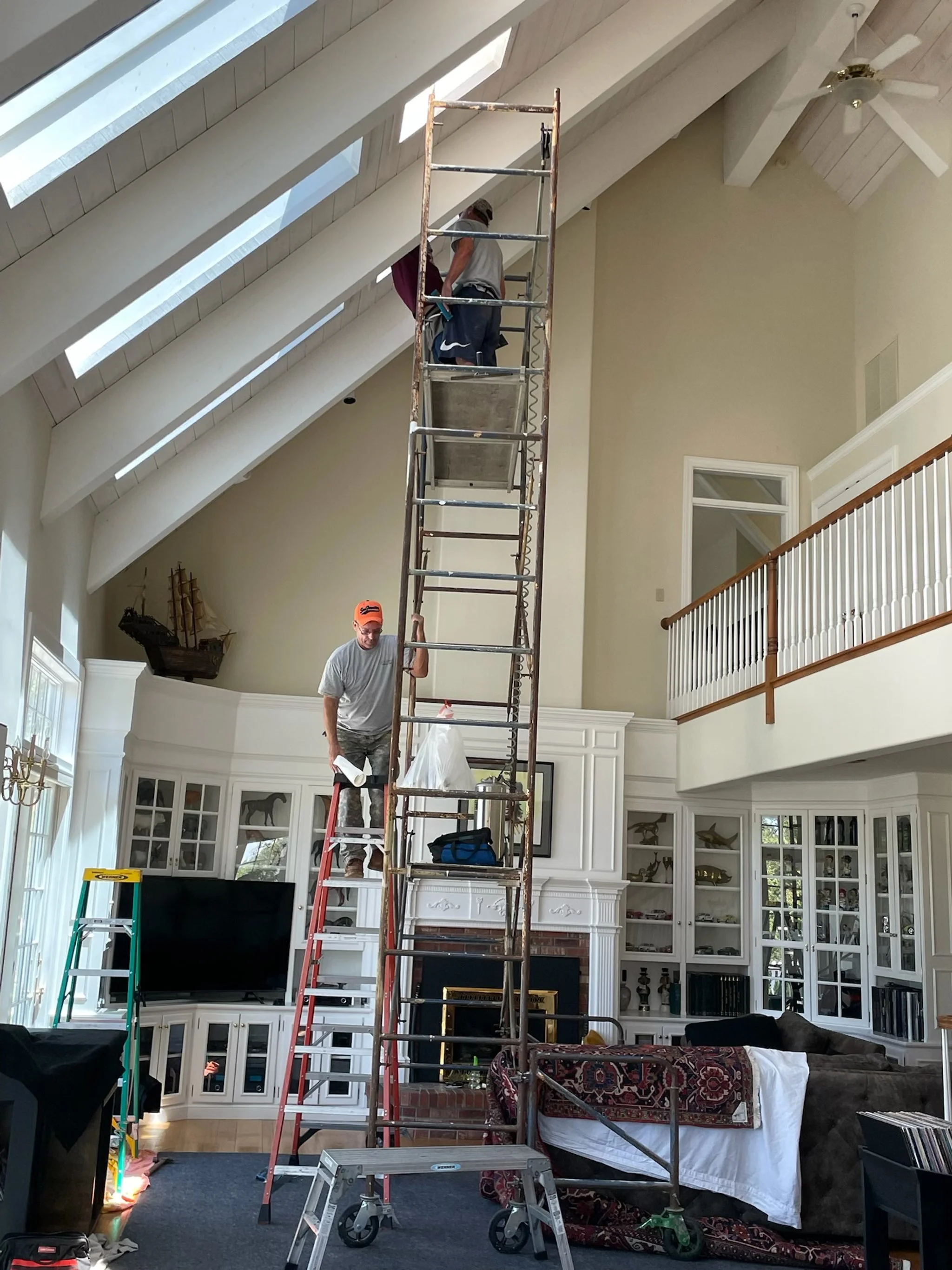Two workers are on different levels of a tall scaffolding inside a living room, possibly installing or repairing something near the ceiling. The living room features a white built-in entertainment center with a large flat-screen TV, white cabinets, a