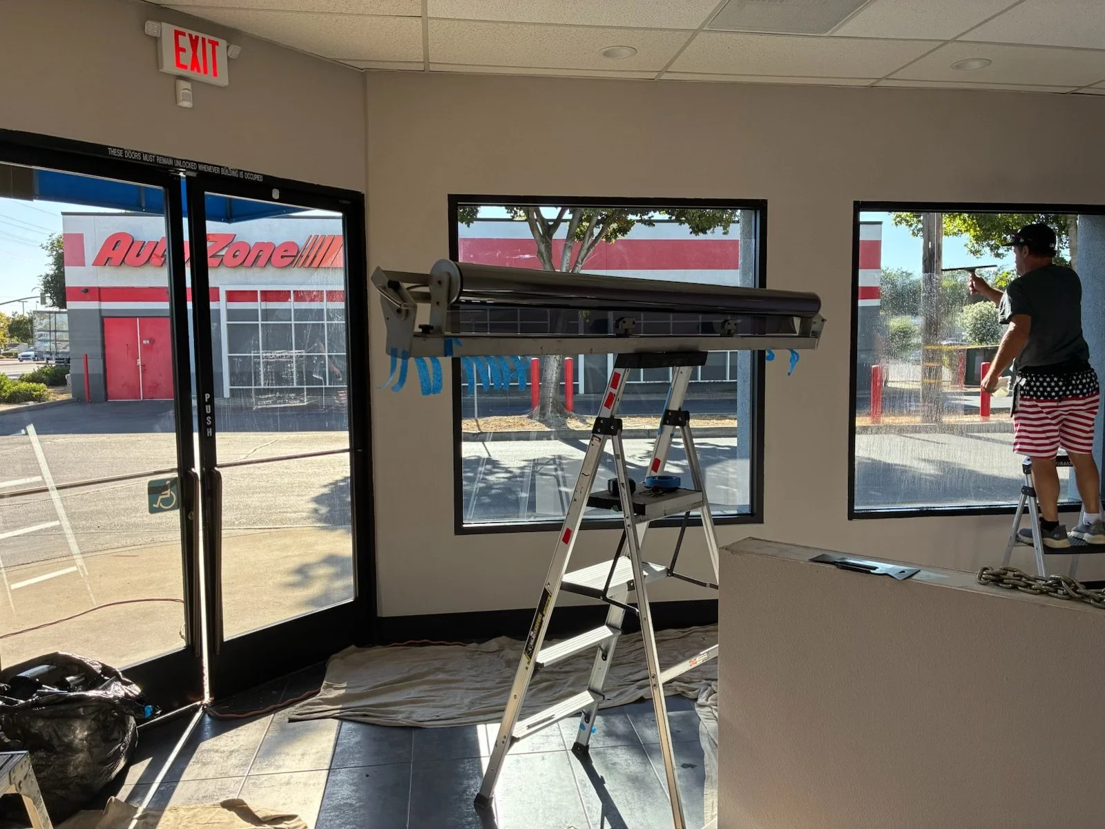 Inside a commercial space with large windows, a ladder and a table near the window, a person working on the window, worker's hands visible on the right, a man in American flag shorts on a small ladder outside, and an AutoZone store visible through th