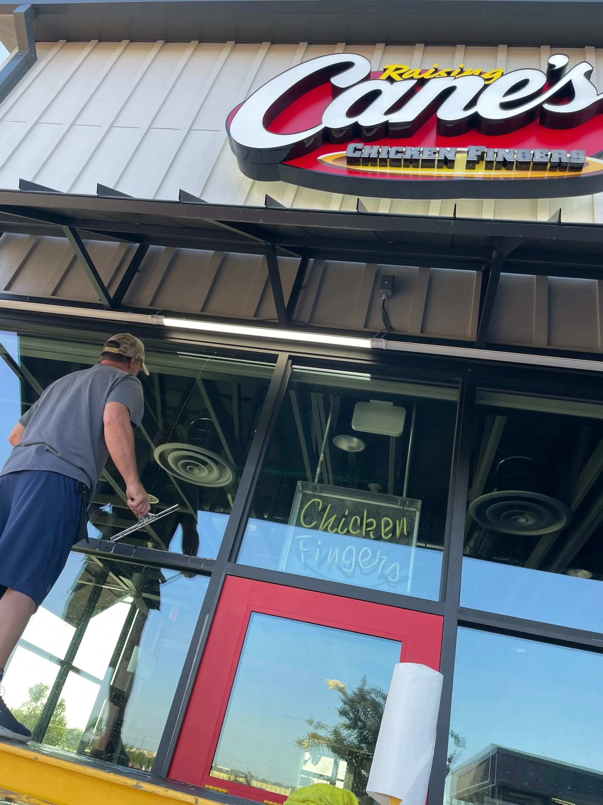 Exterior of Raising Cane's restaurant with a sign reading 'Raising Cane's Chicken Fingers' and a person cleaning the window.