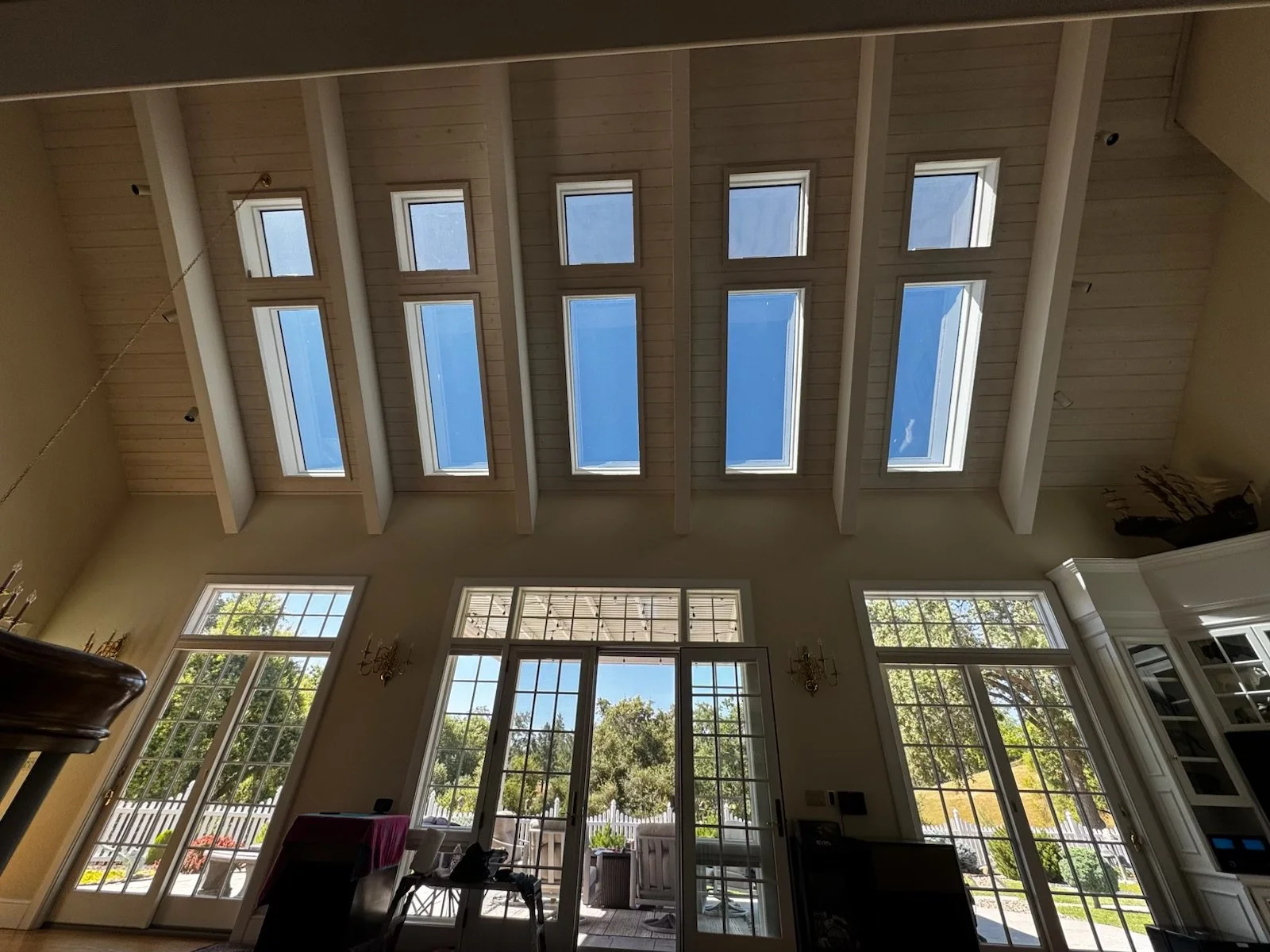 Interior view of a spacious room with large windows and skylights, high ceiling with exposed beams, and a view of the outdoors with trees and a patio.
