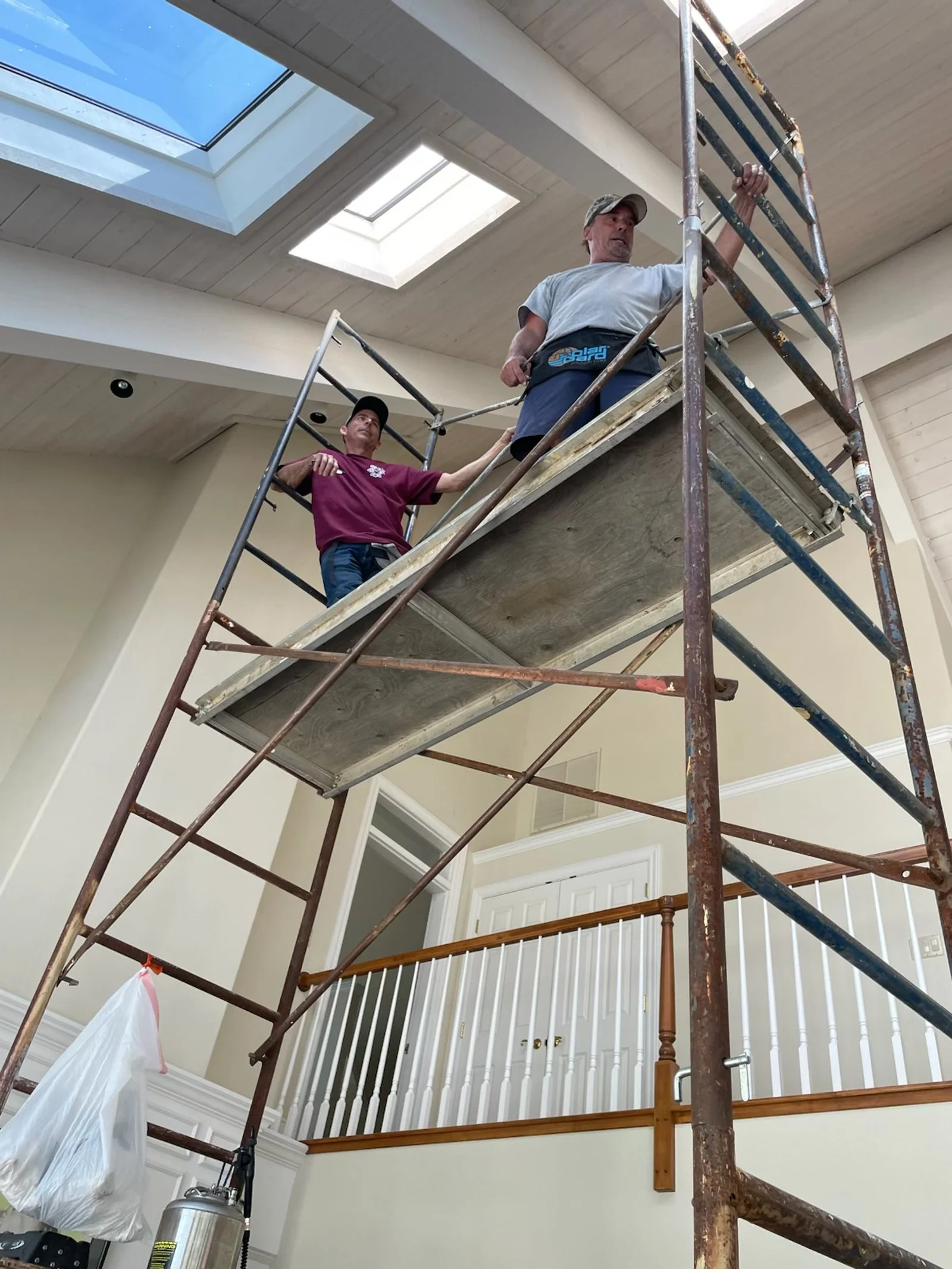 Two workers on scaffolding inside a house, working on the ceiling. The ceiling has skylights, and the house has a high ceiling with a skylight window.