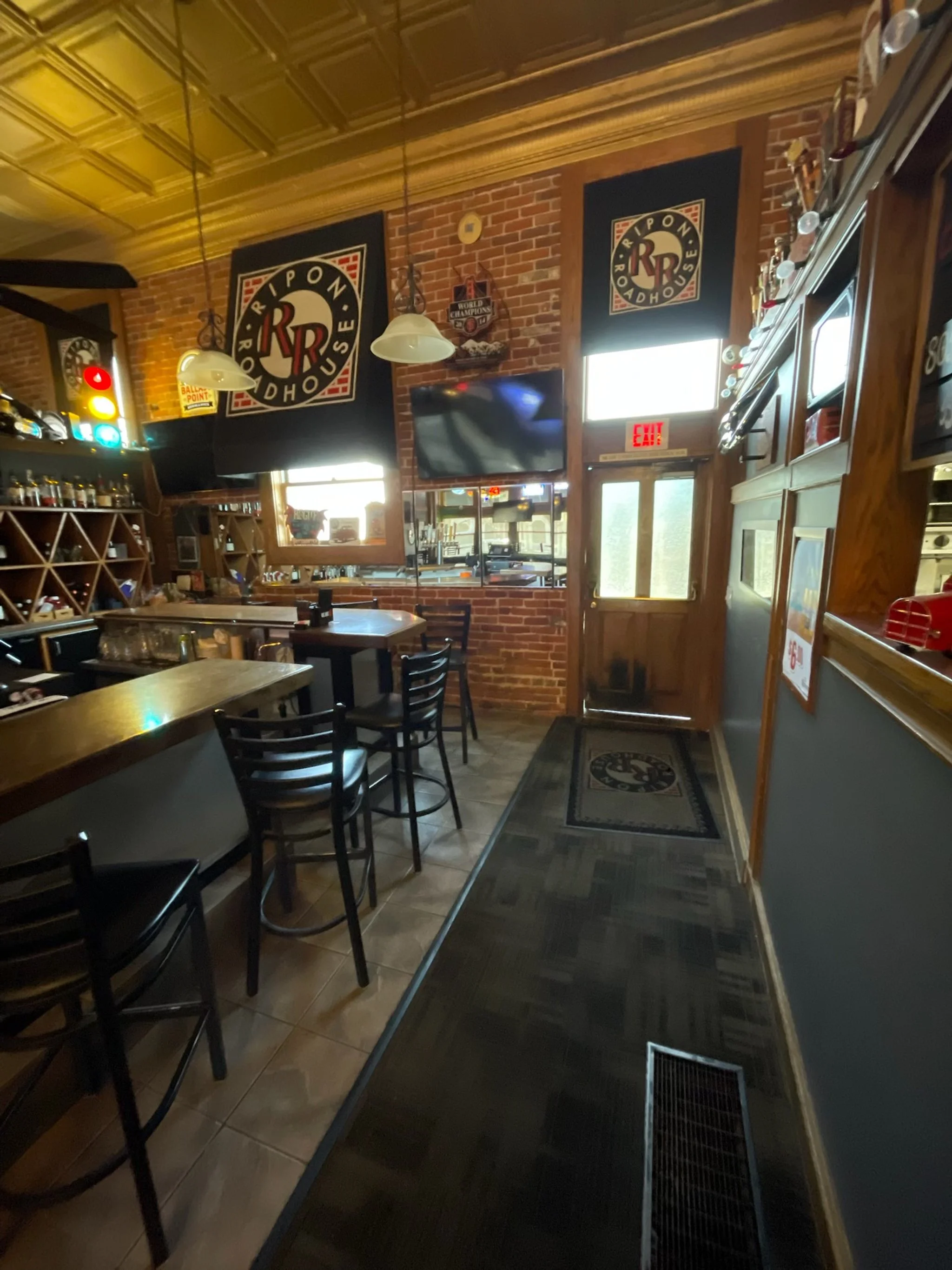Inside a pub with brick walls, wooden ceiling, and bar stools. Decorative signs and a large RPM Roadhouse sign are on the walls. There is a TV on the wall and sunlight coming through the windows.