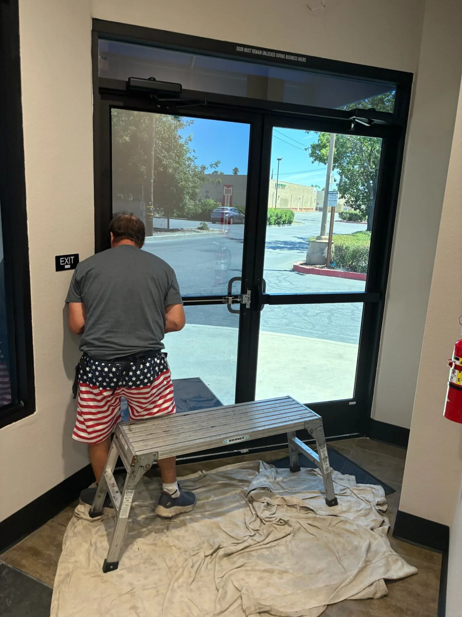 A man in patriotic shorts and a gray shirt is working on a glass door inside a building, standing on a ladder on a dirty cloth, with the outdoors visible through the door.
