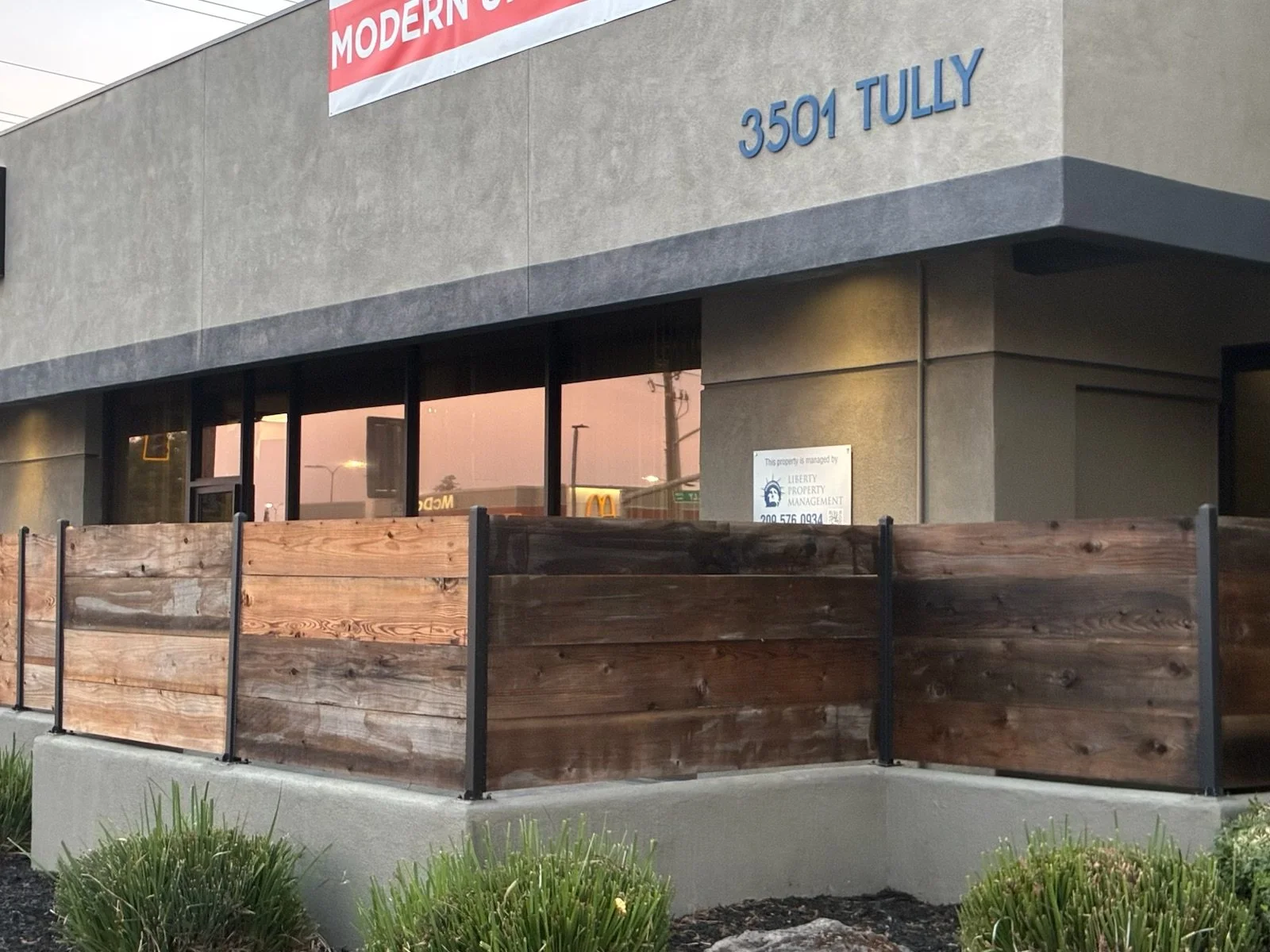 Commercial building with large windows and a sign indicating the address 3501 Tully. The front has a wooden fence and some green plants.