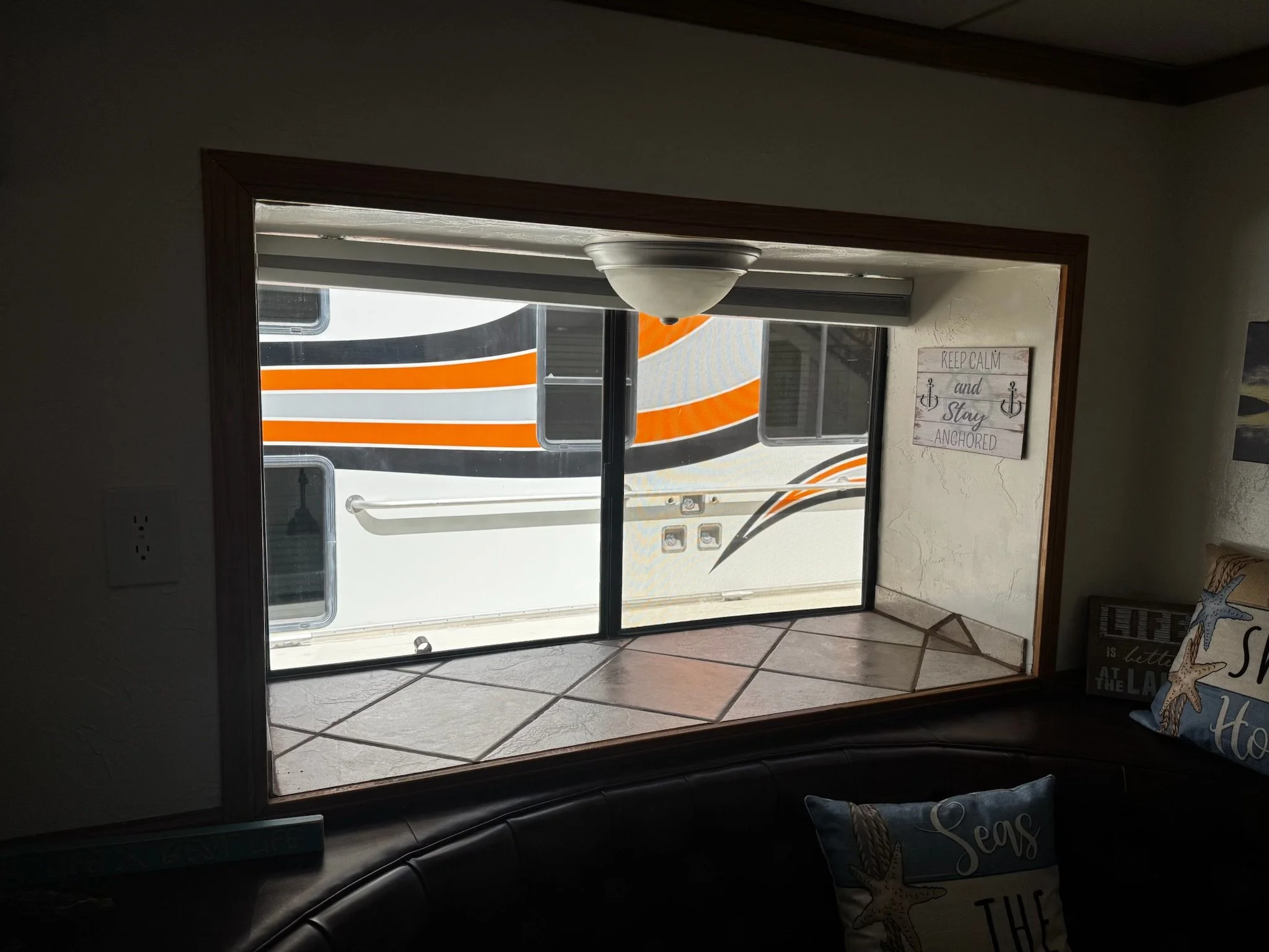 View of an RV through a window from inside a room with beige walls, tropical-themed cushions, and a sign that reads "Keep Calm and Stay Anchored."
