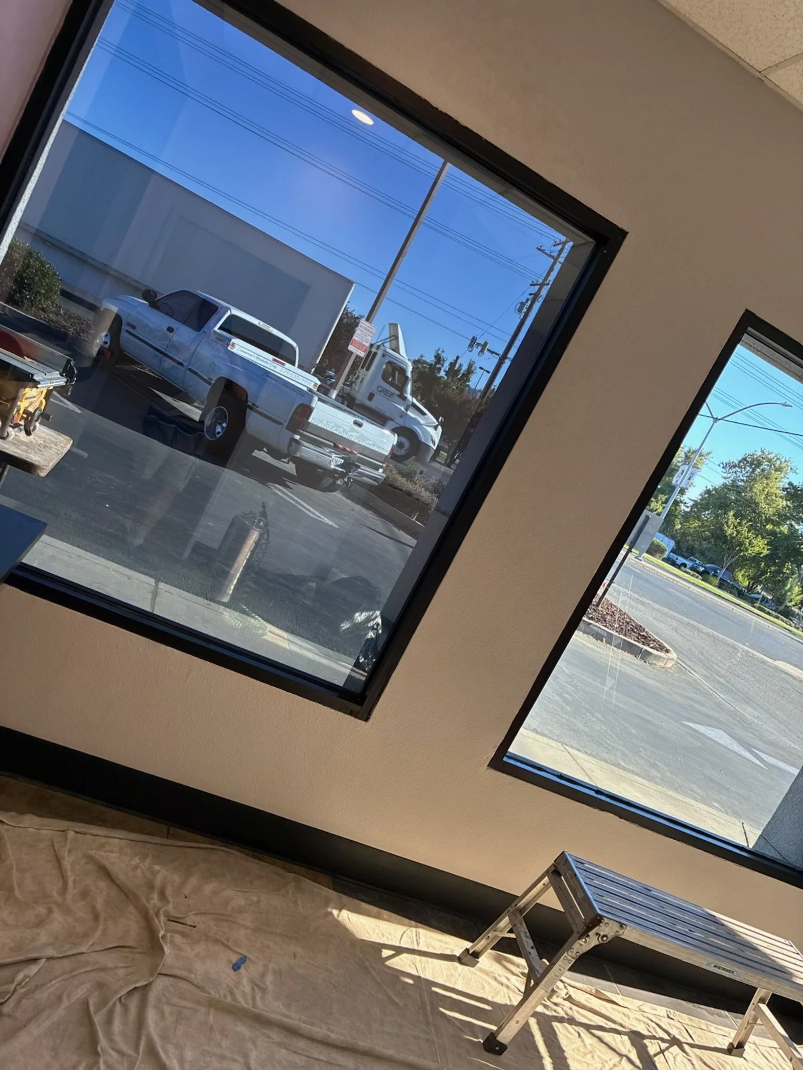 Photo of the interior of a building with large windows, showing a parking lot outside with a white pickup truck and a white delivery truck. The floor is covered with a cloth, and there is a small metal step ladder in the corner.