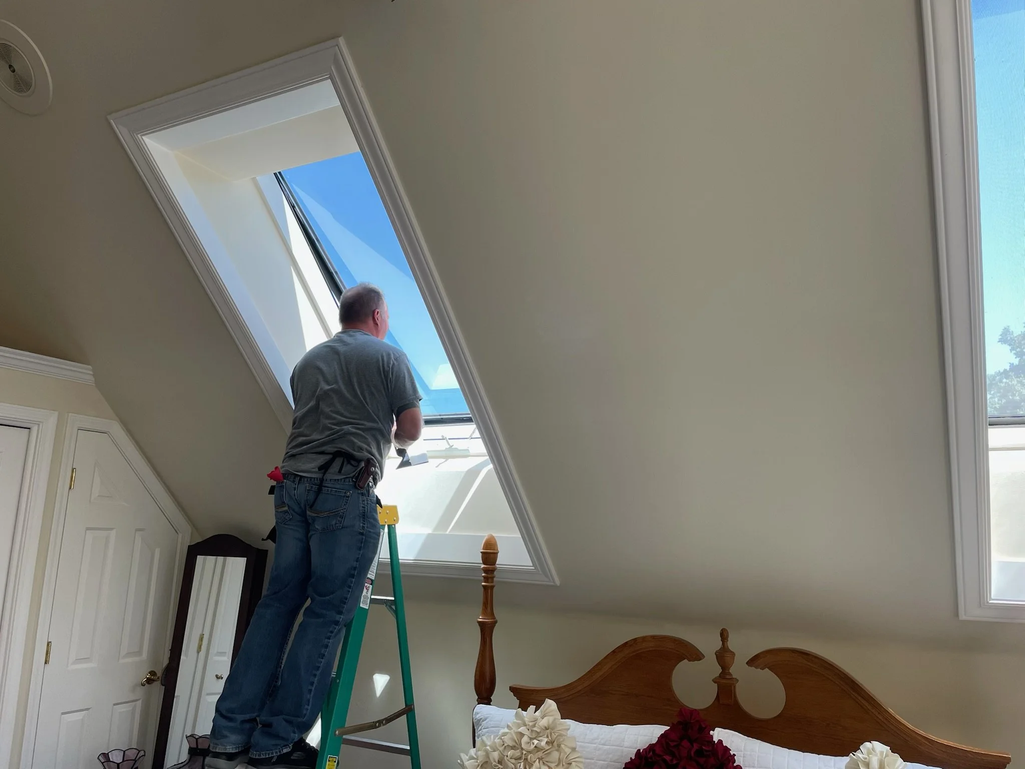 A man standing on a ladder installing or cleaning a large skylight window in a sloped attic ceiling, with a bed and floral pillows visible below.