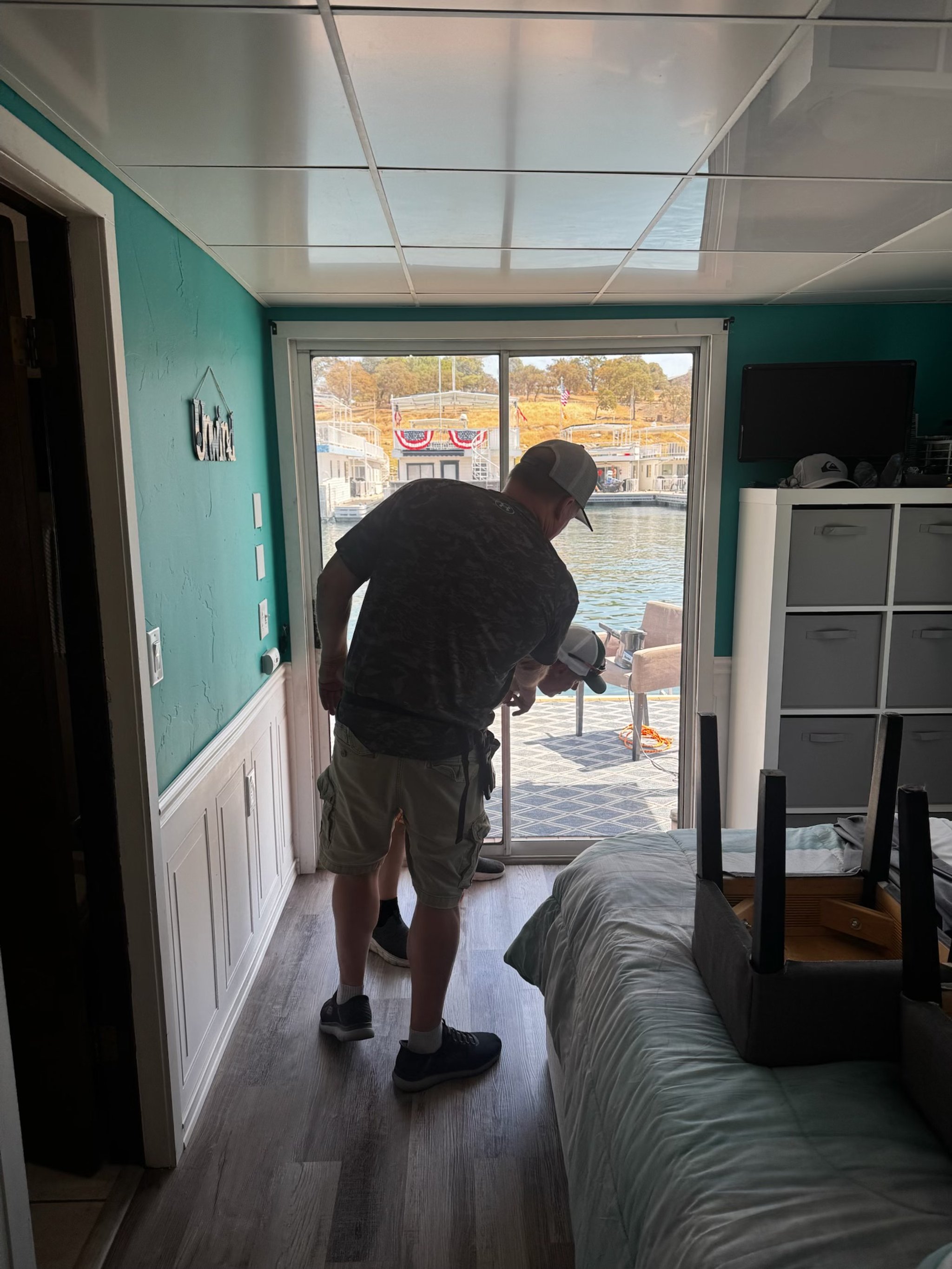 A person with a baseball cap vacuuming the floor inside a room by a large glass sliding door with a view of a waterway and outdoor furniture.