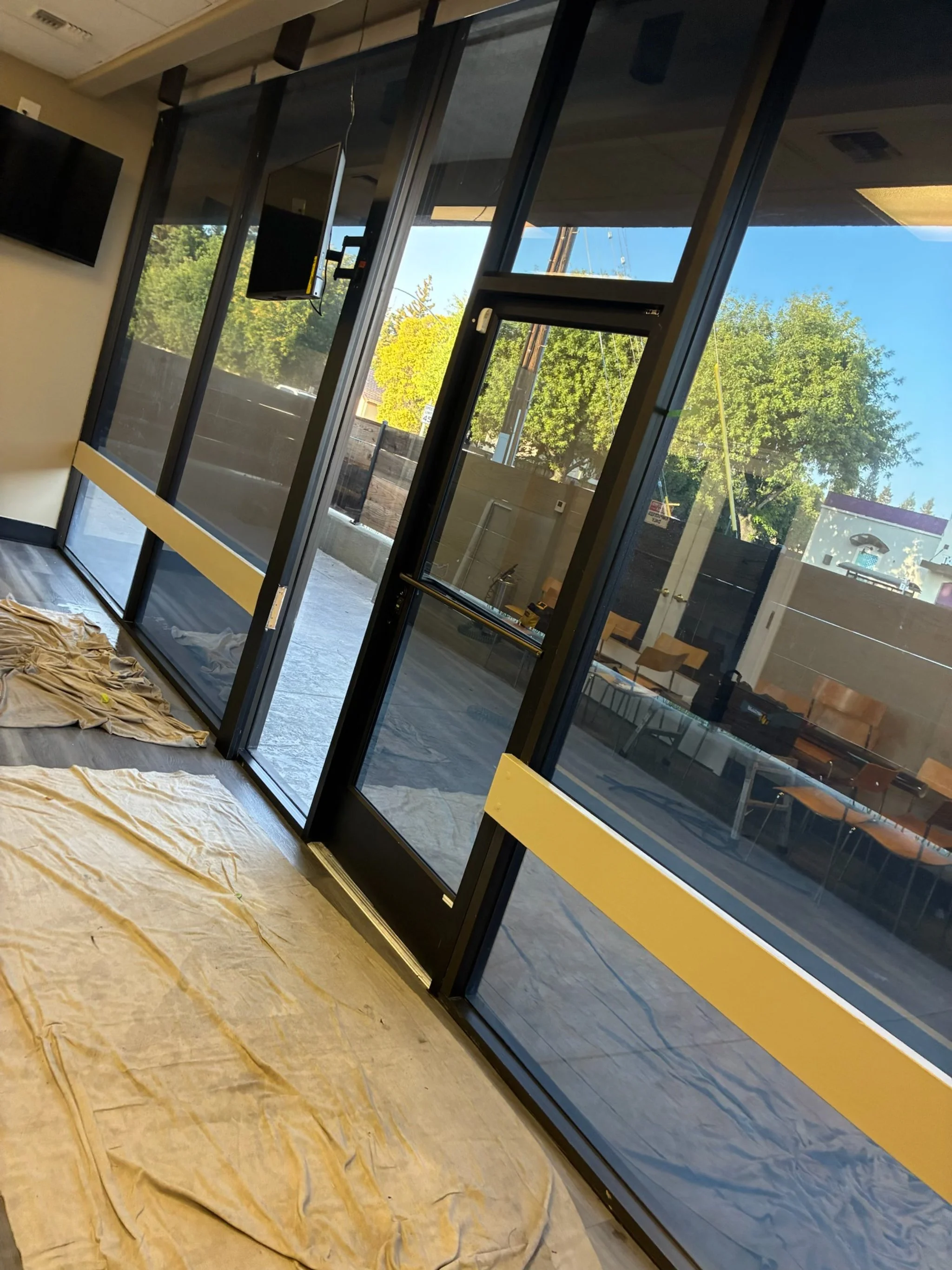 The image shows an indoor space with large glass windows and doors, some covered with protective plastic or paper on the floor. Outside, there are trees with green leaves and a bright blue sky.