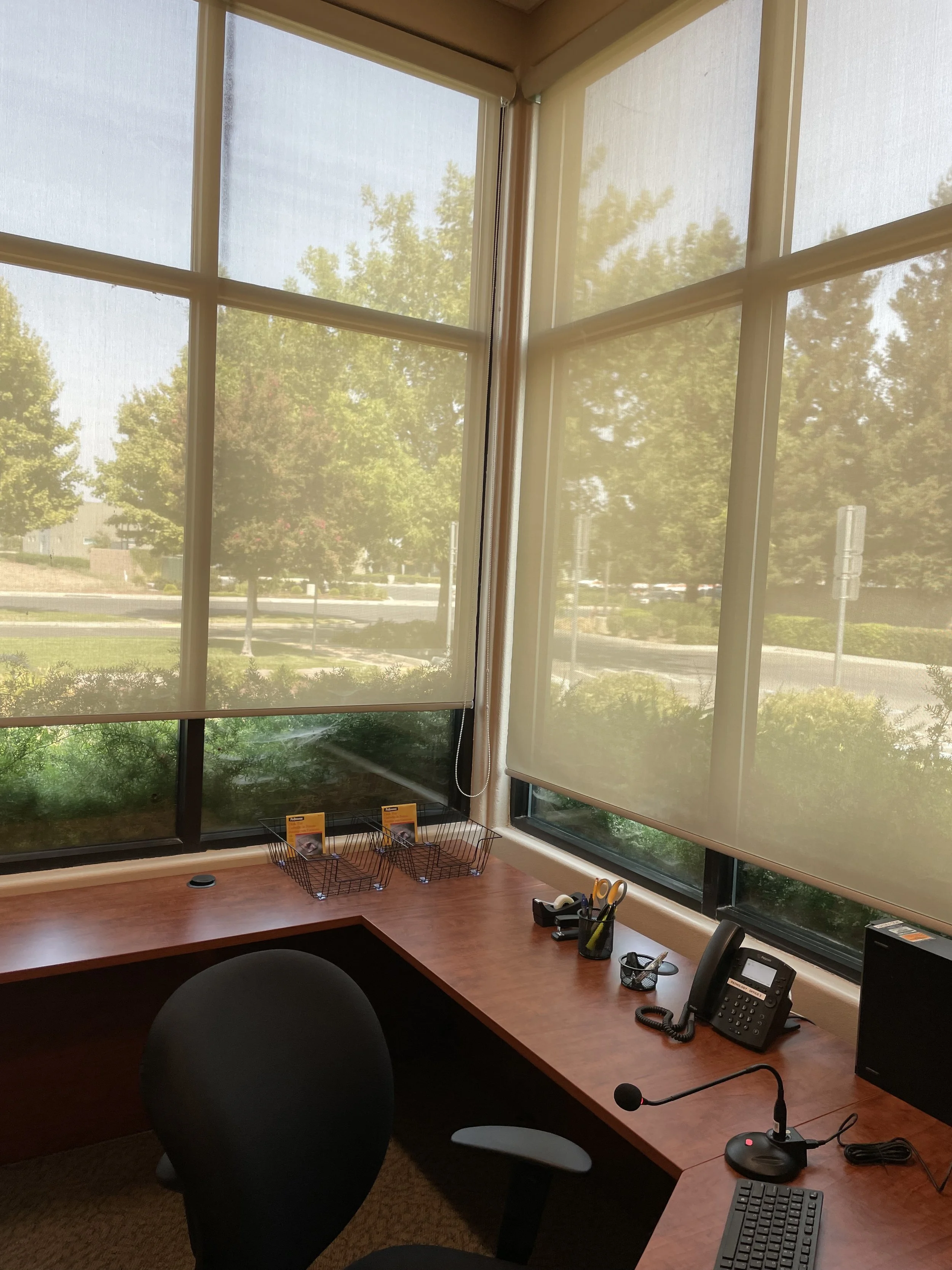 Office with a large window covered by beige roller blinds, showing trees and a parking lot outside.