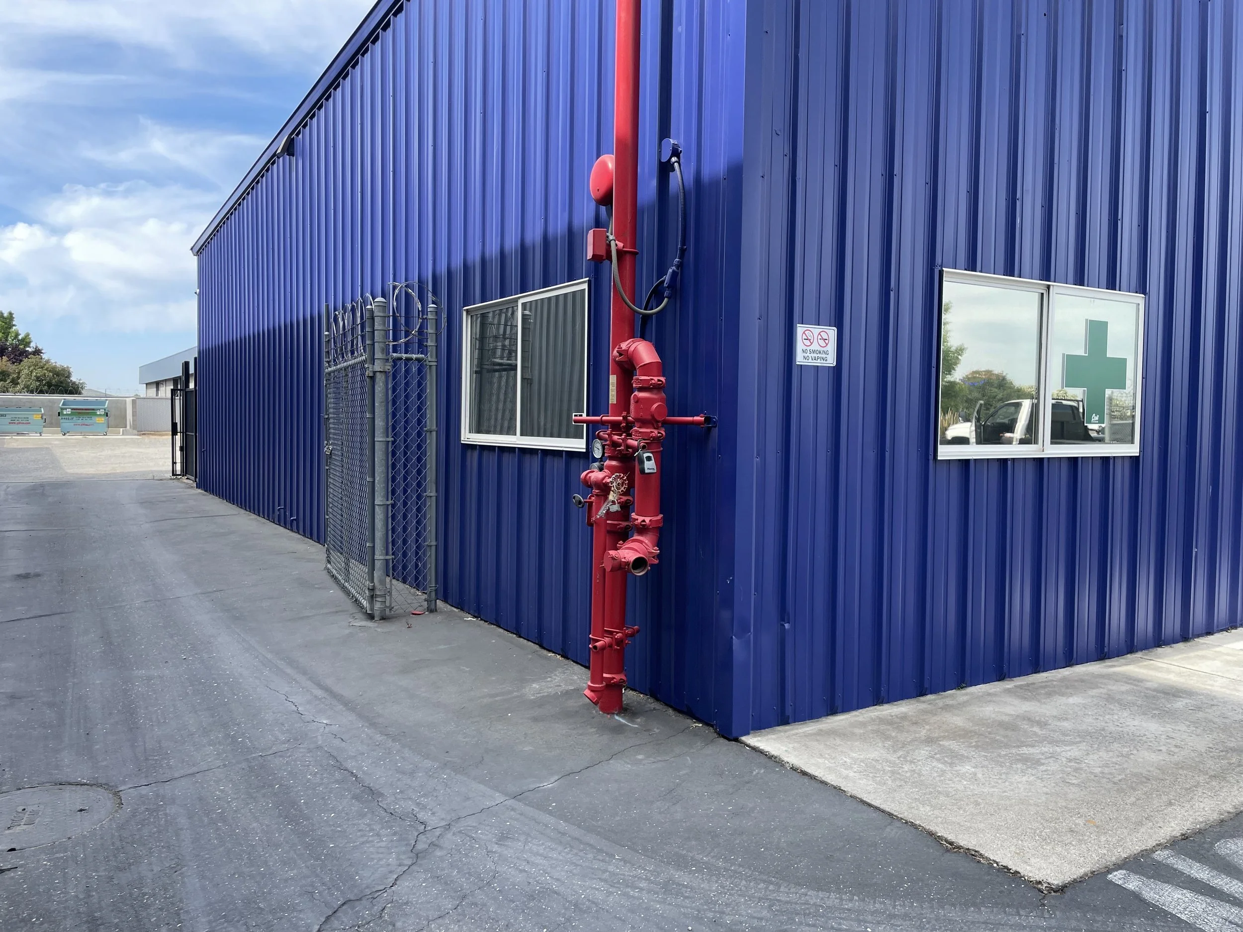 Exterior of a blue metal building with two windows, a red fire hydrant pipe, and a 'No Smoking' sign on the wall.