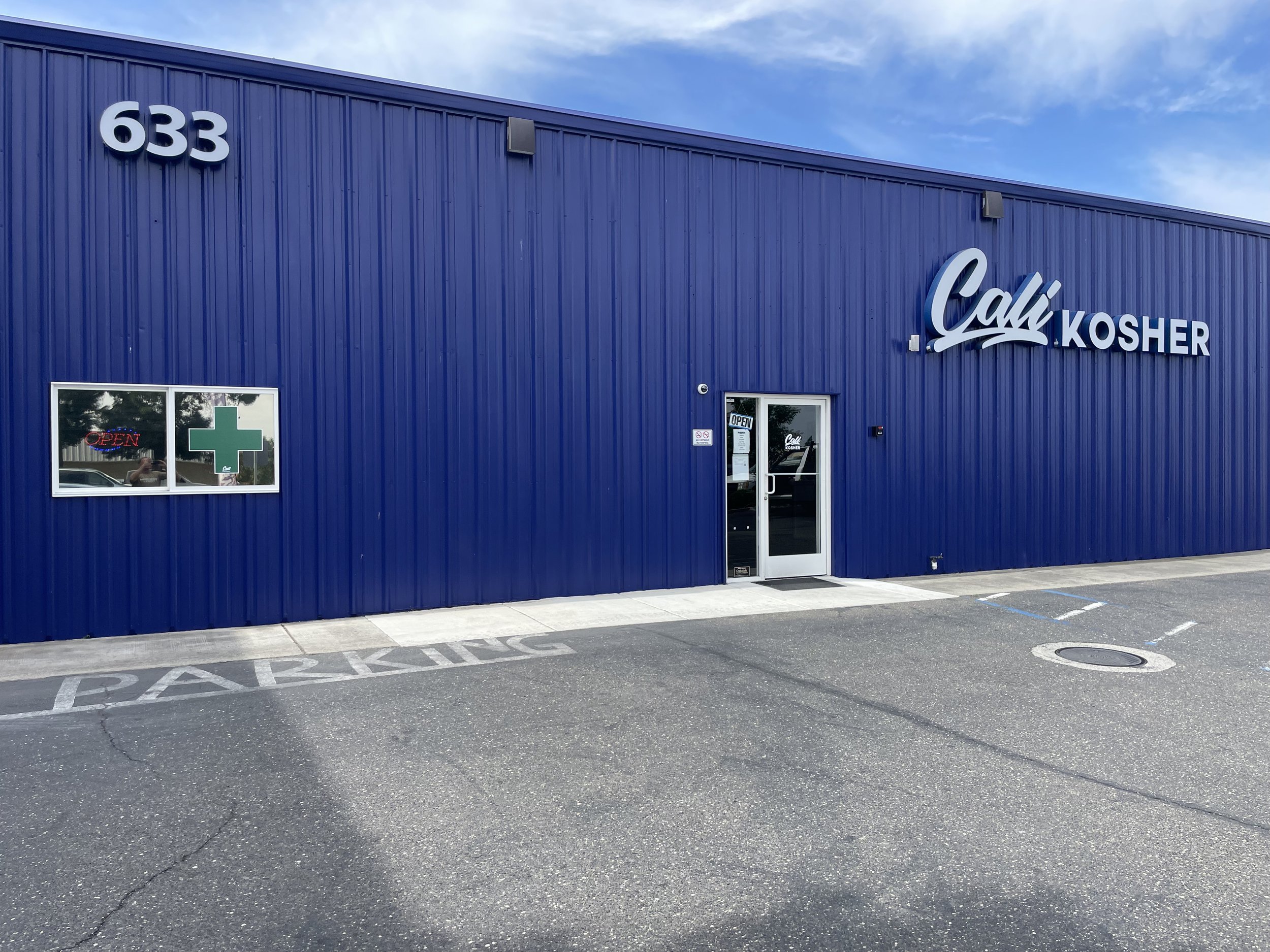 Blue building with the sign 'Cali Kosher' and the number 633 on it, a window with an 'Open' sign and a green cross, and a parking space marked 'Parking' in front.