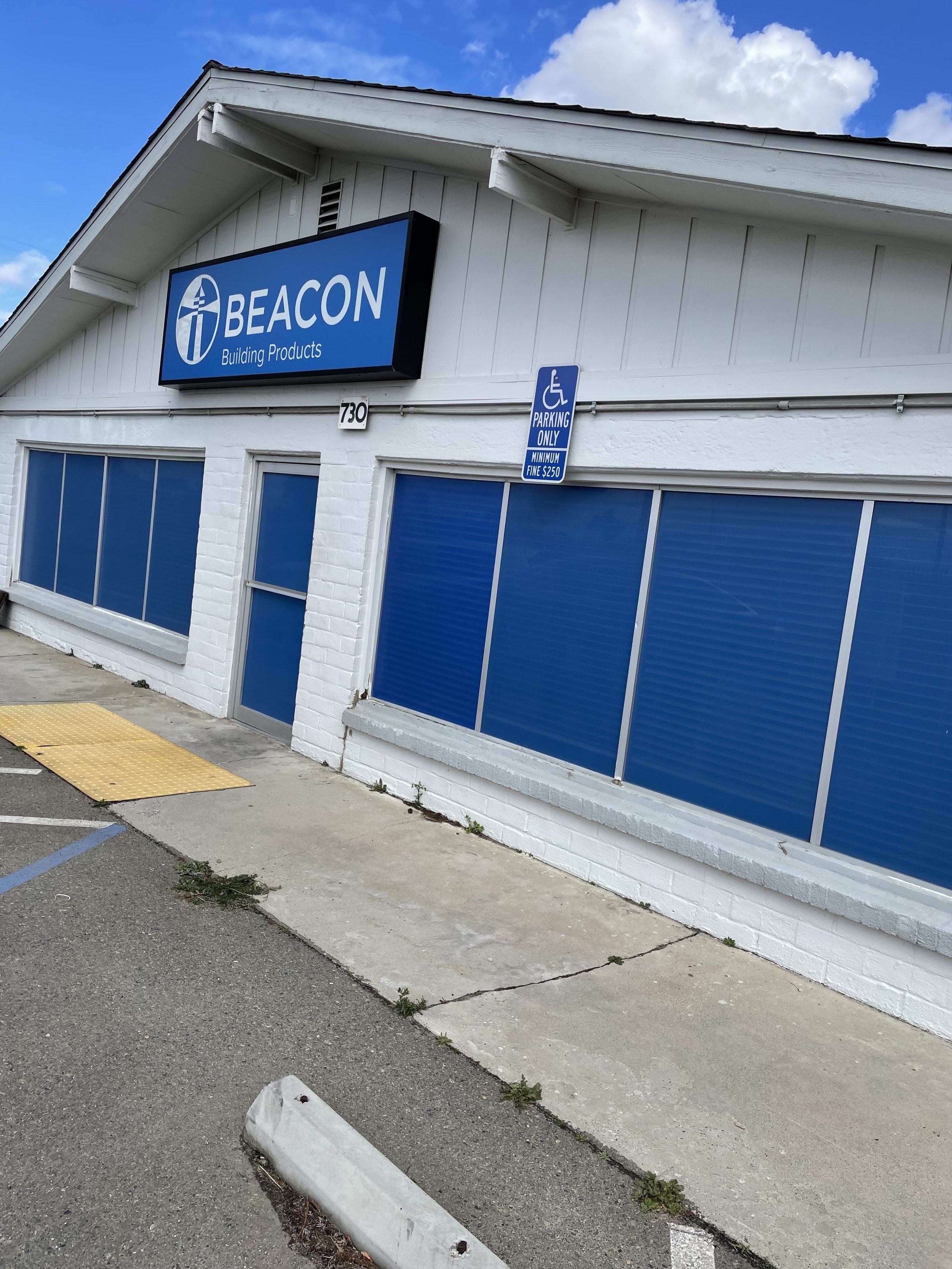 Storefront building with a blue sign that reads 'BEACON Building Products,' a handicap parking sign, a white brick exterior, and blue window shades.