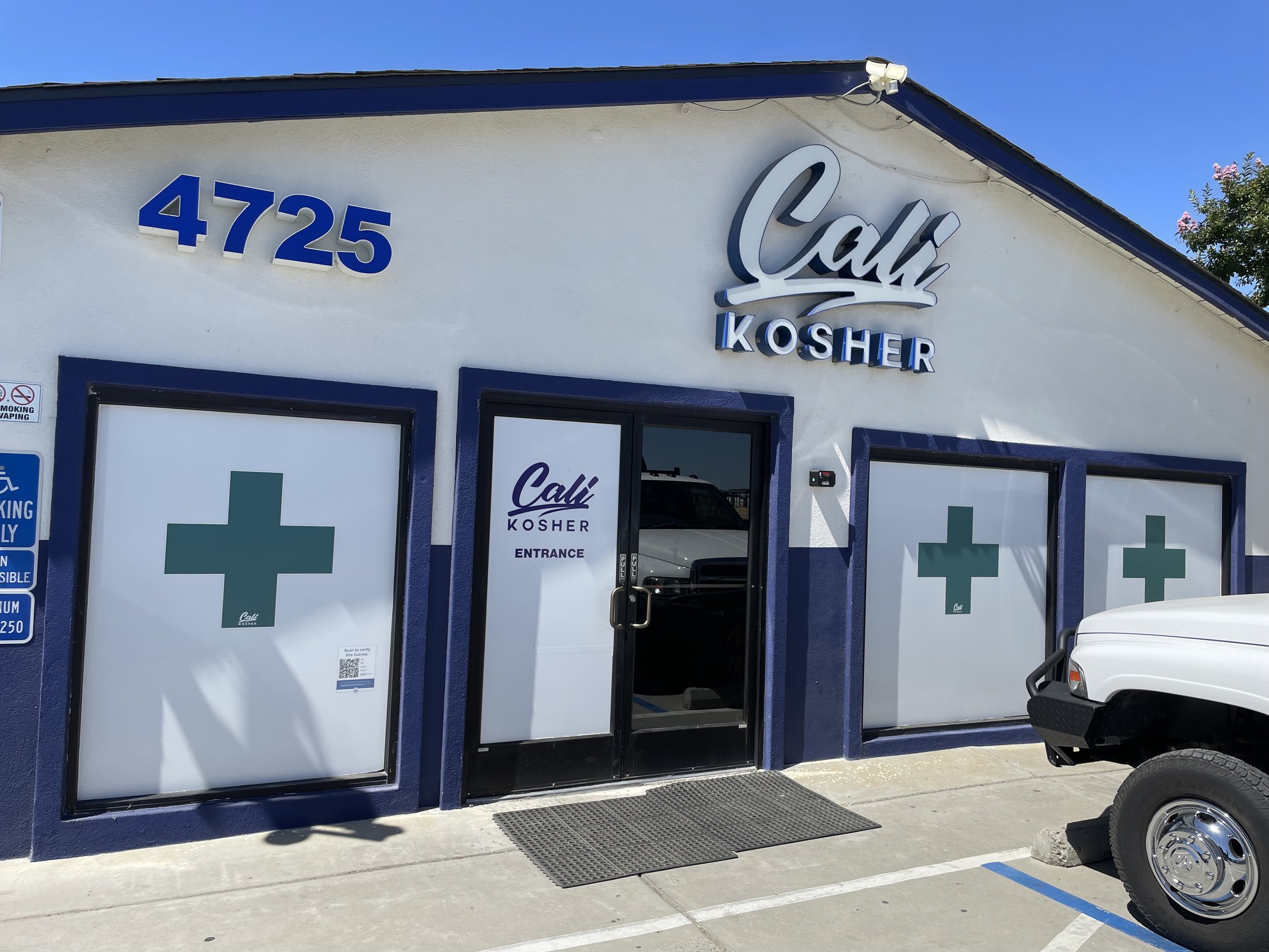 Exterior view of Cali Kosher pharmacy with the address 4725, featuring signs with green cross symbols on windows, a white building with blue accents, and a white vehicle parked outside.