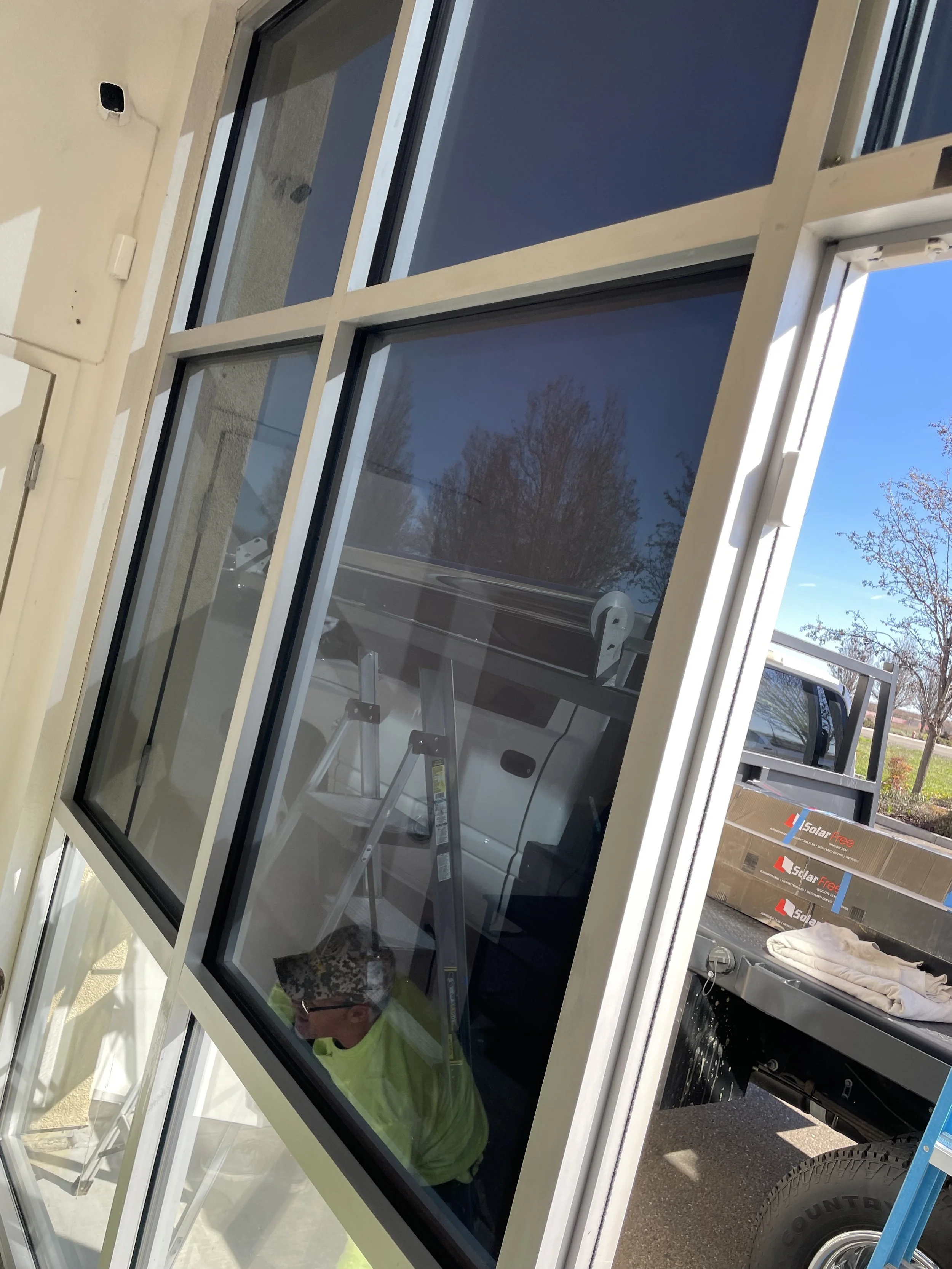Worker installing or inspecting a large window on the side of a building, with a ladder and a truck nearby.
