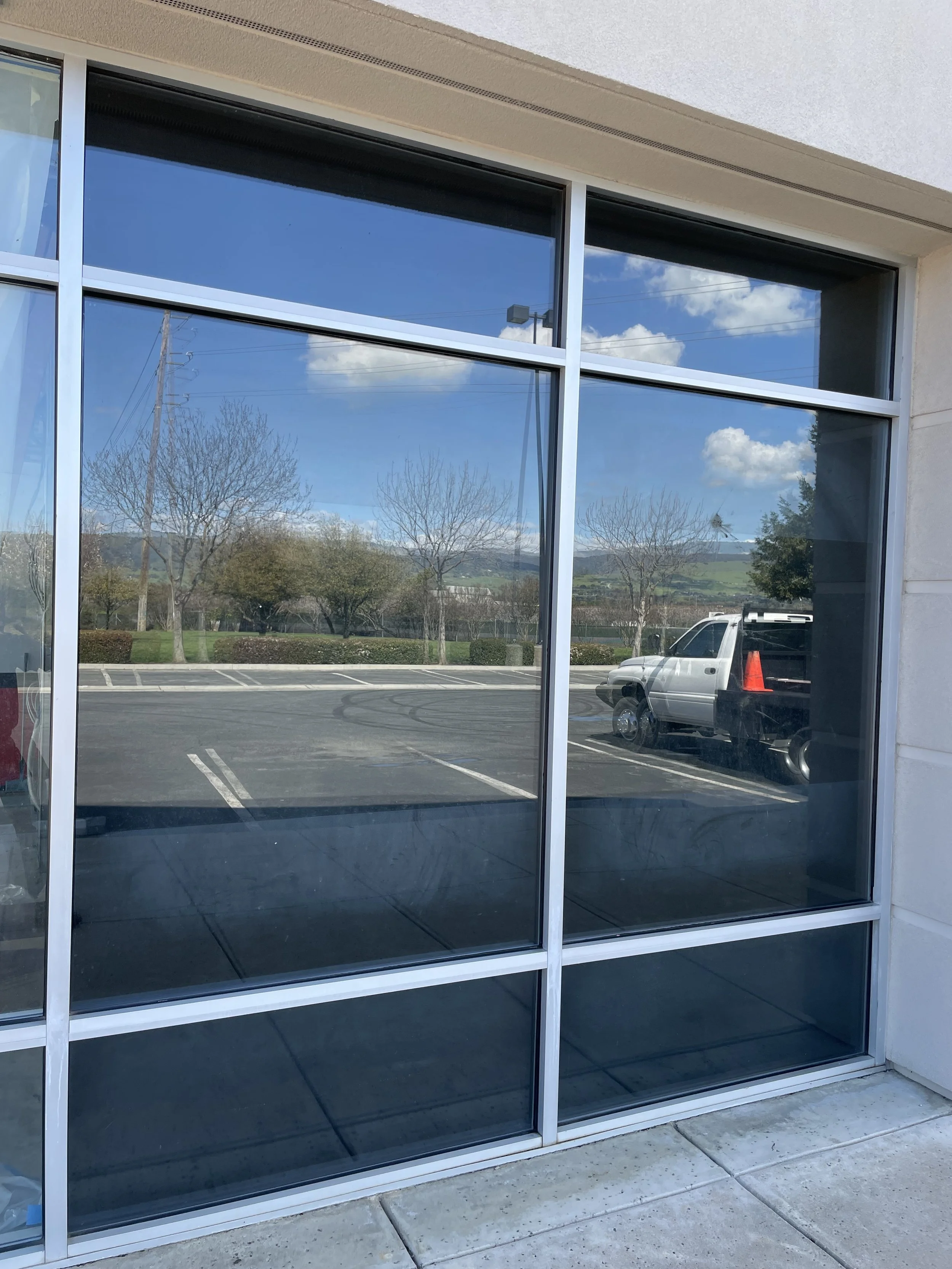 Reflection of the parking lot, trees, and blue sky with clouds seen through a large glass window of a building.