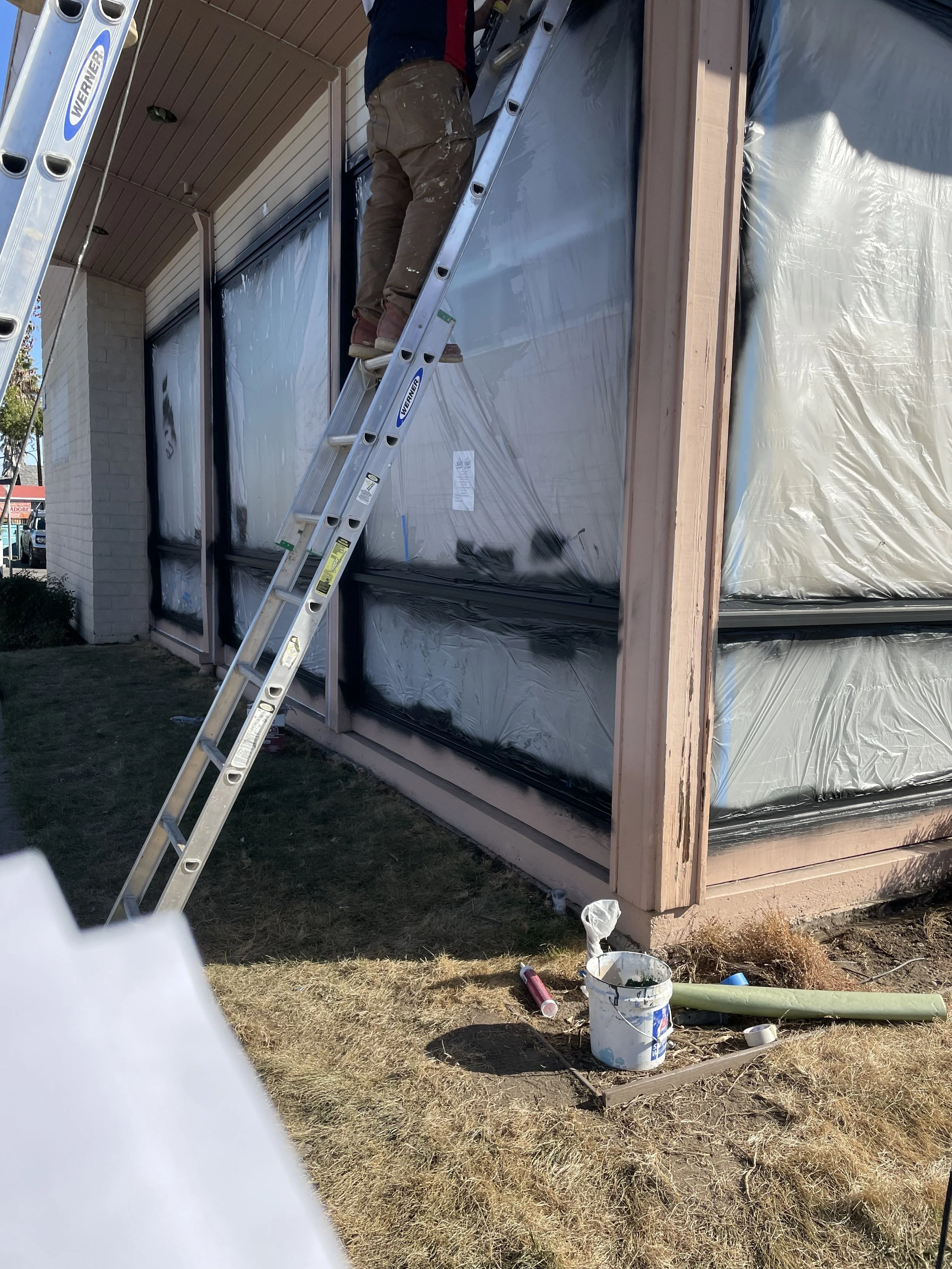 A person standing on a ladder working on the exterior wooden frame of a building under construction, with plastic sheeting covering windows, and open paint cans and tools on the ground nearby.