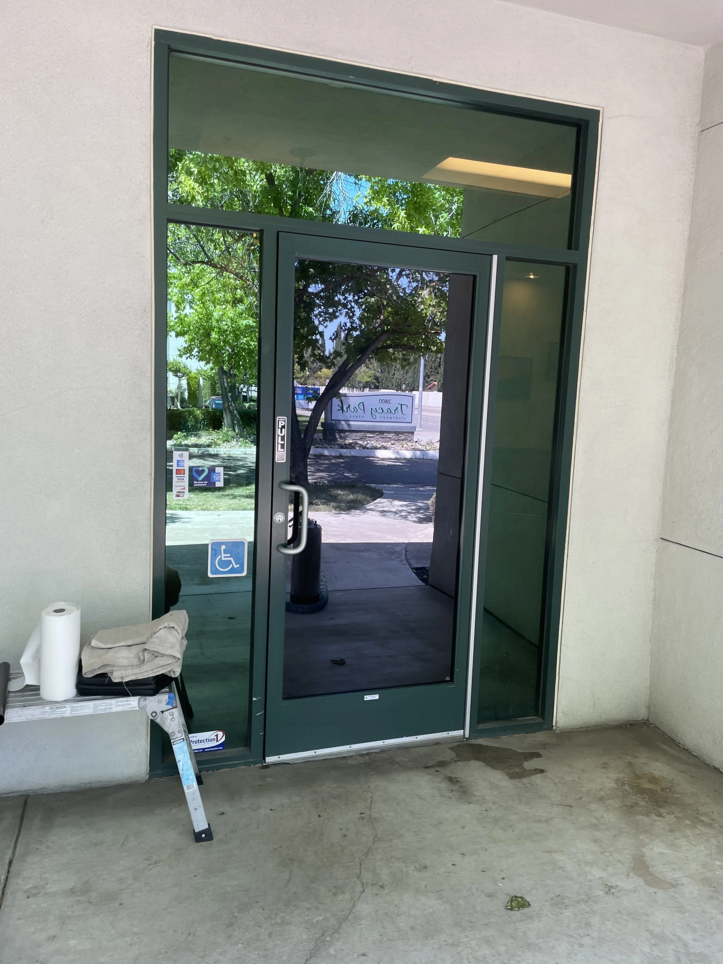 Glass door with handicap accessibility sticker, outside greenery visible through the glass, and a small table with paper towels and cloth inside.