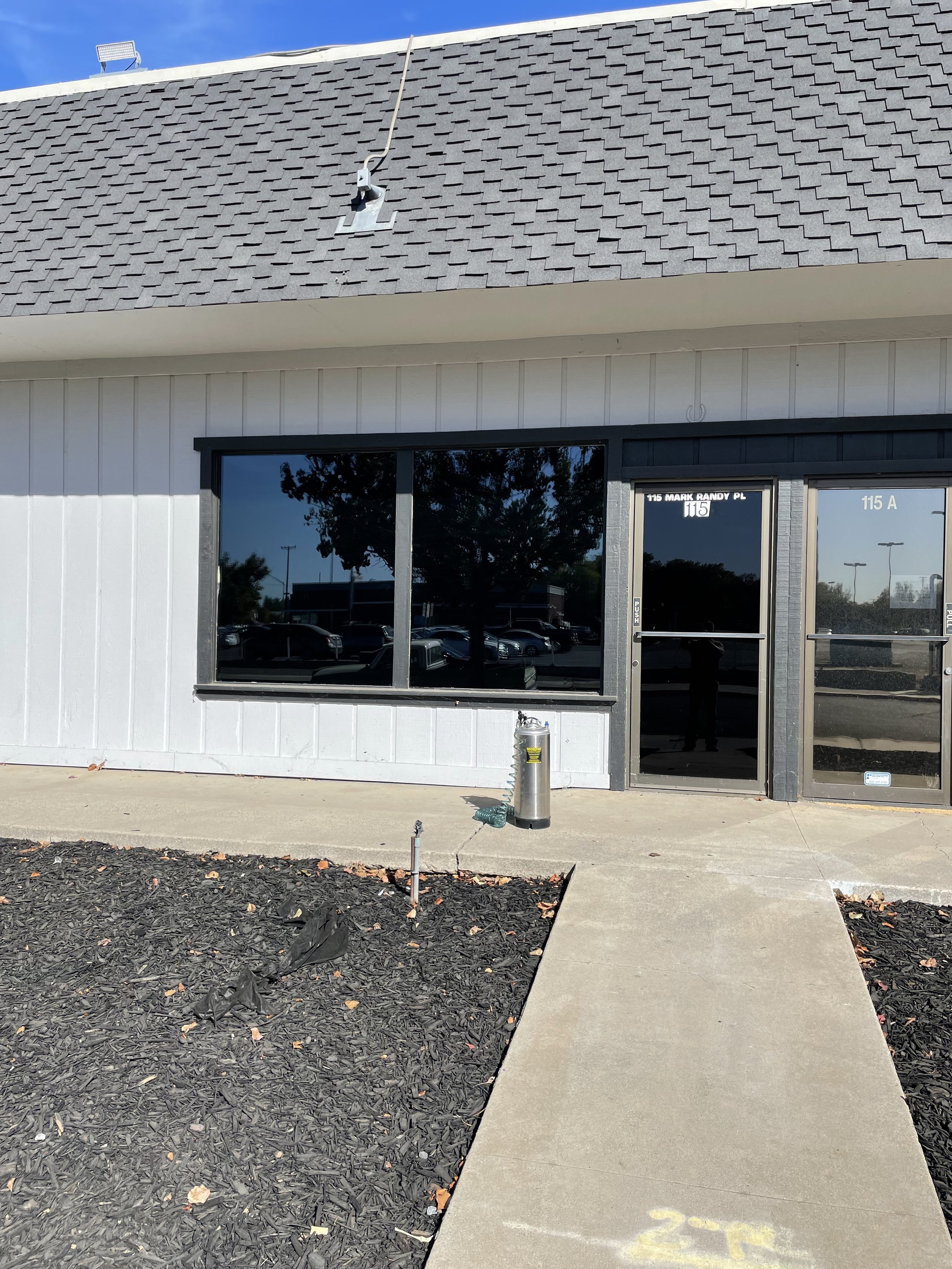 Exterior view of a commercial building with black-framed windows and a glass door, gray shingled roof, a sidewalk leading to the entrance, and a parking lot reflected in the windows. A fire hydrant is in front of the window, and some landscaping with