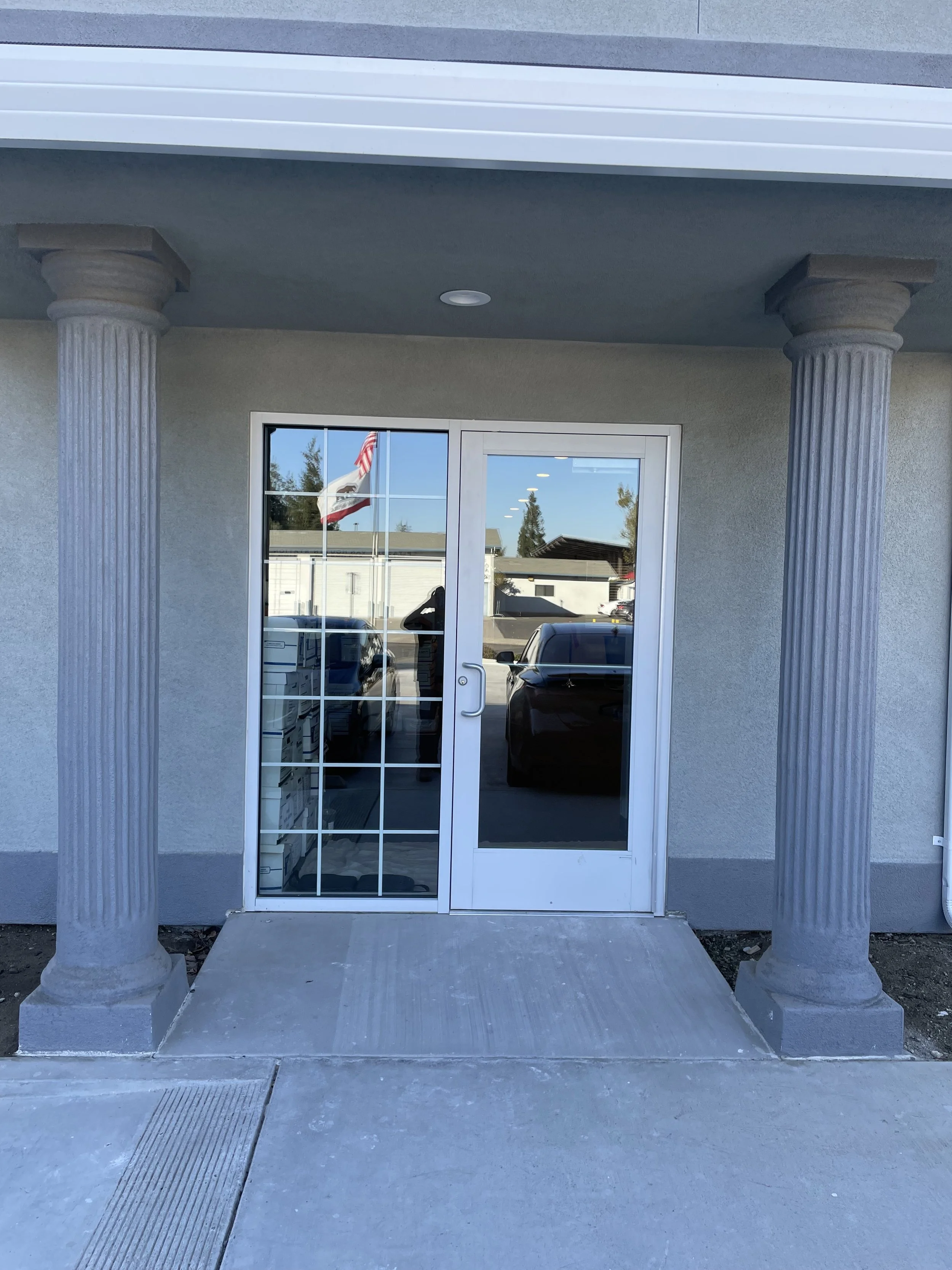 Glass door with white frame, flanked by two classical-style stone columns, leading into a building. Reflection of parked cars and a flagpole with the American and California flags in the glass door.