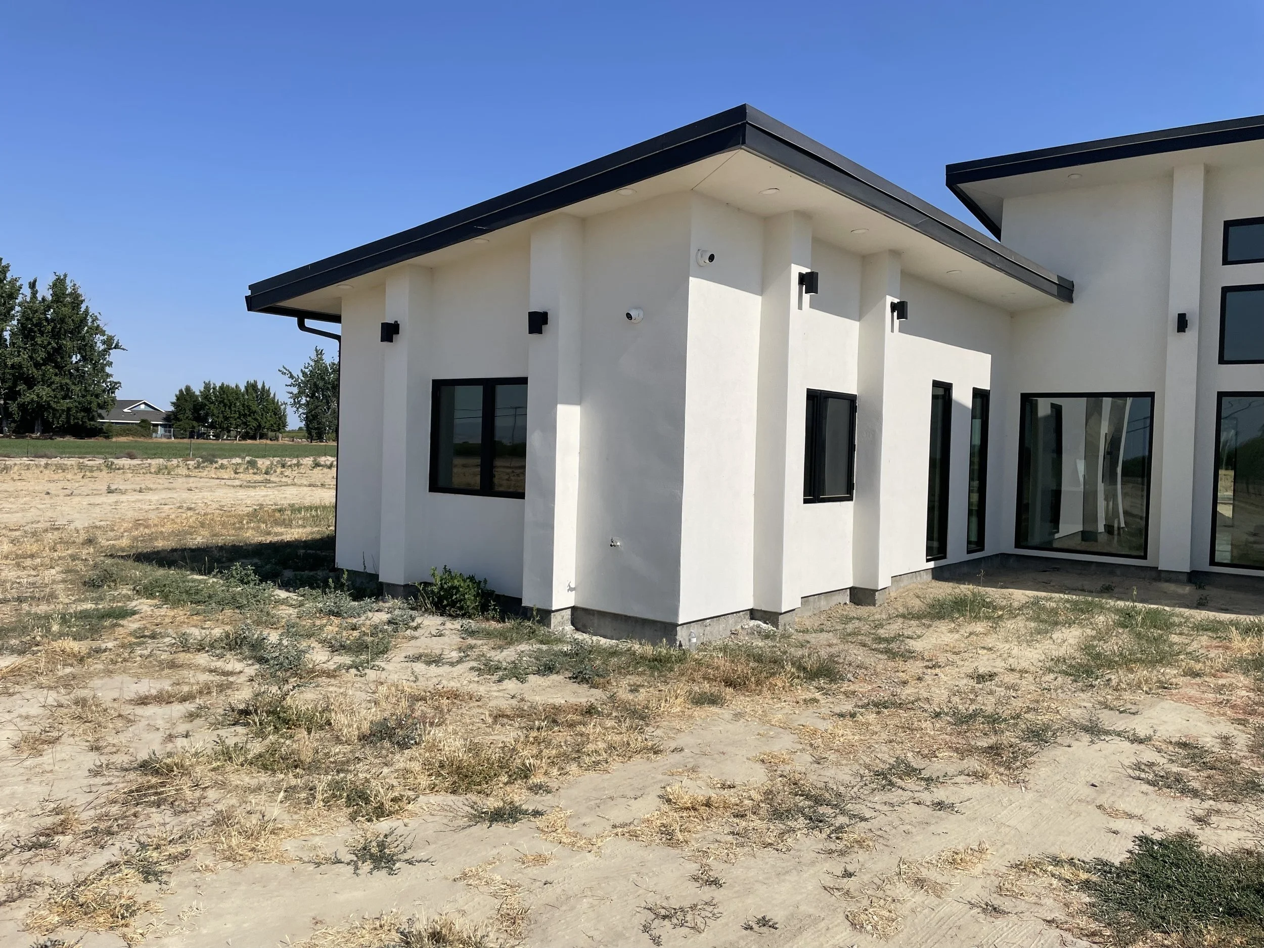 Modern white house with black window frames and a flat roof, situated in a dry, sparsely vegetated area with a blue sky overhead.