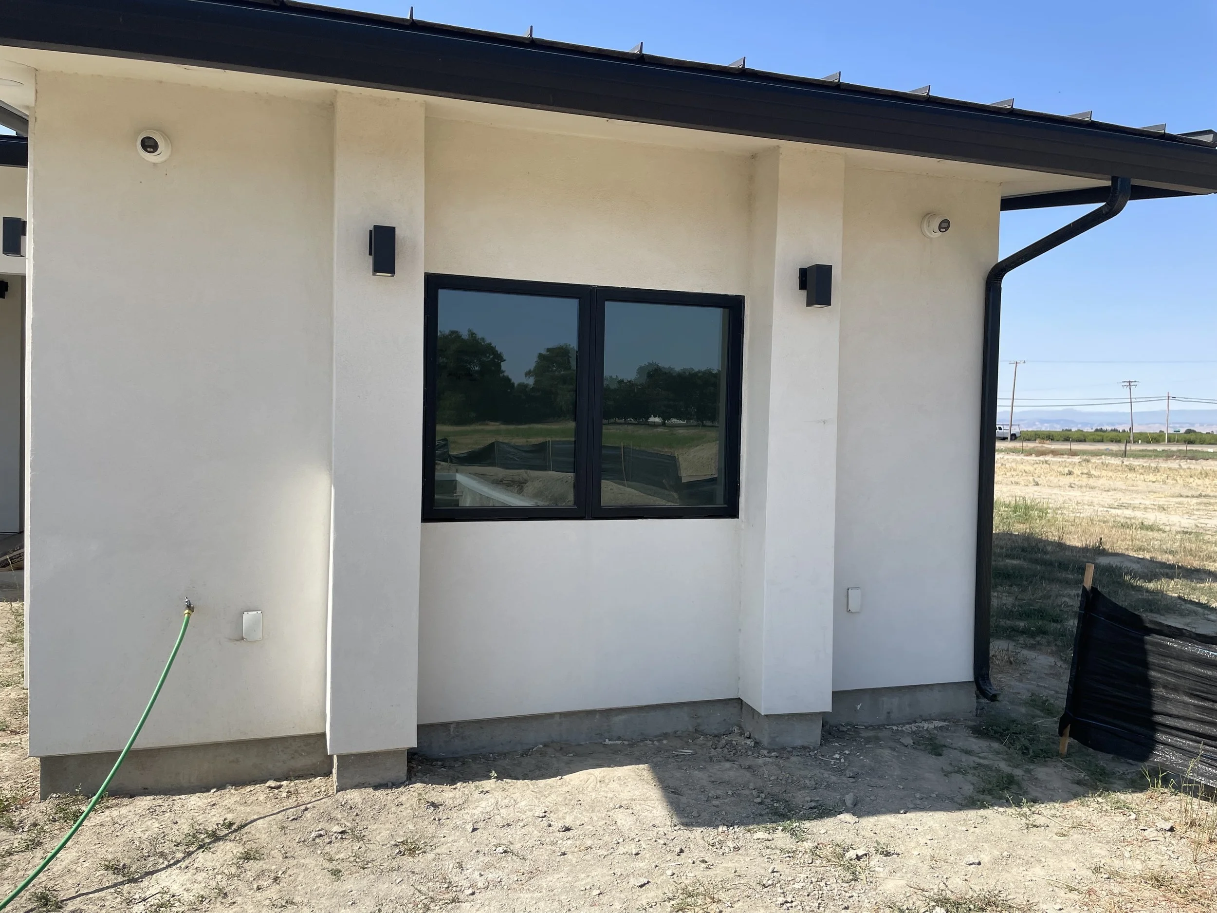The exterior of a modern house with white stucco walls, a large window, black wall lights, and a black gutter, set in an area with dirt ground and a clear blue sky.