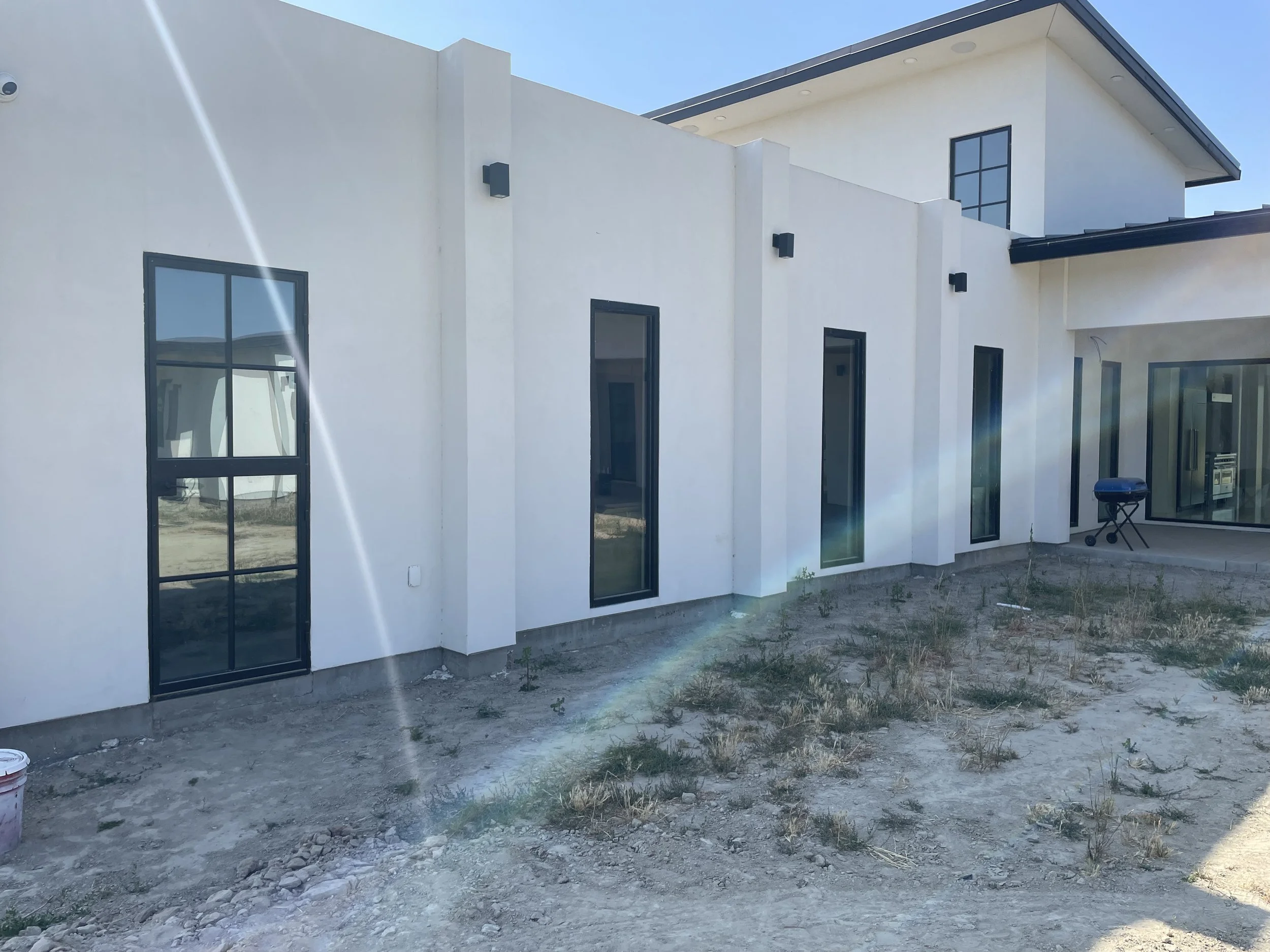 Modern white house with black framed windows and sliding glass doors, construction site with dirt and sparse grass in the yard, a small barbecue grill, clear blue sky, and sunlight creating glare.