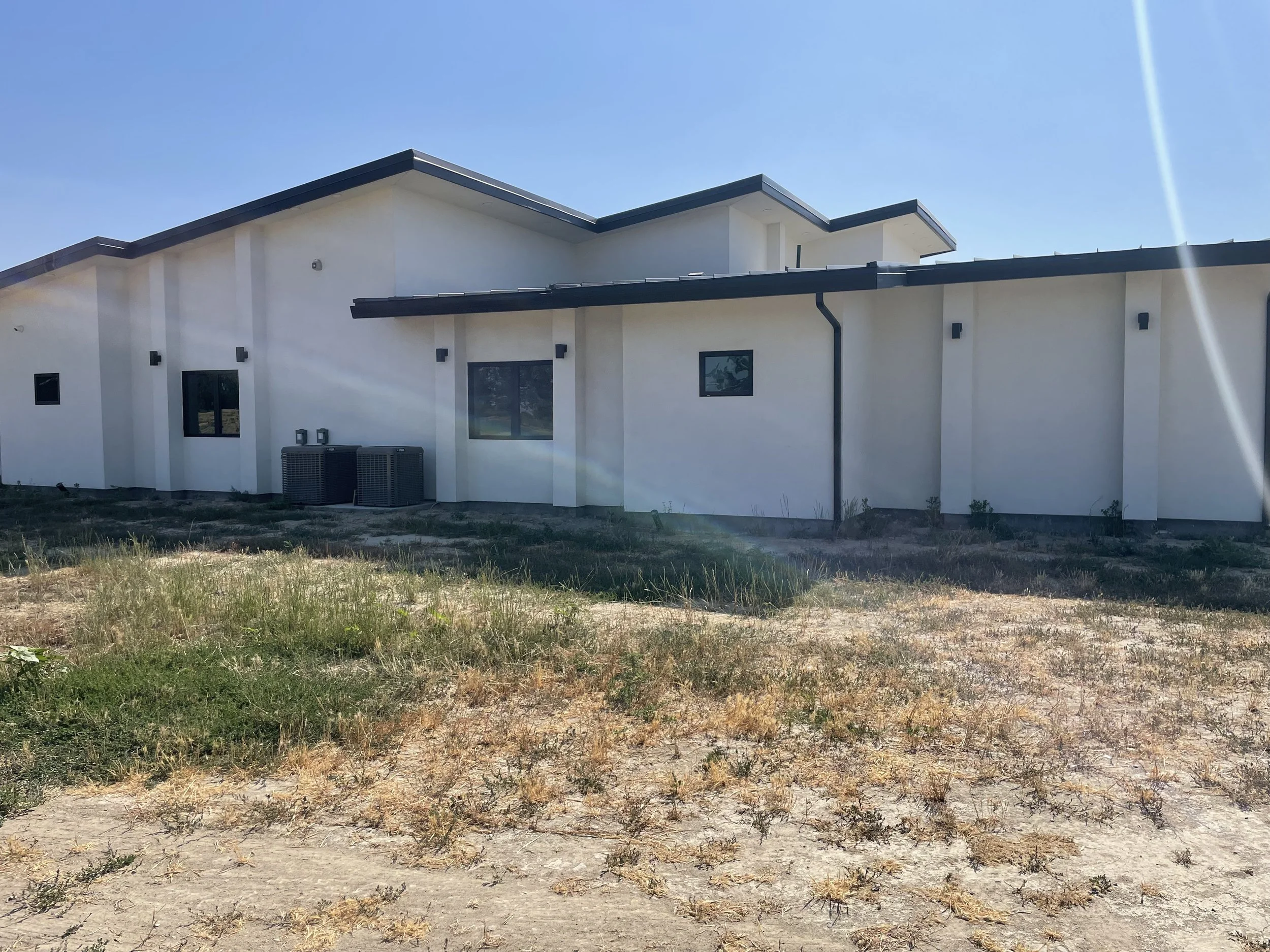 A modern, single-story white house with a flat, sloped roof on a sunny day, with air conditioning units outside and a patchy, dry lawn in the foreground.