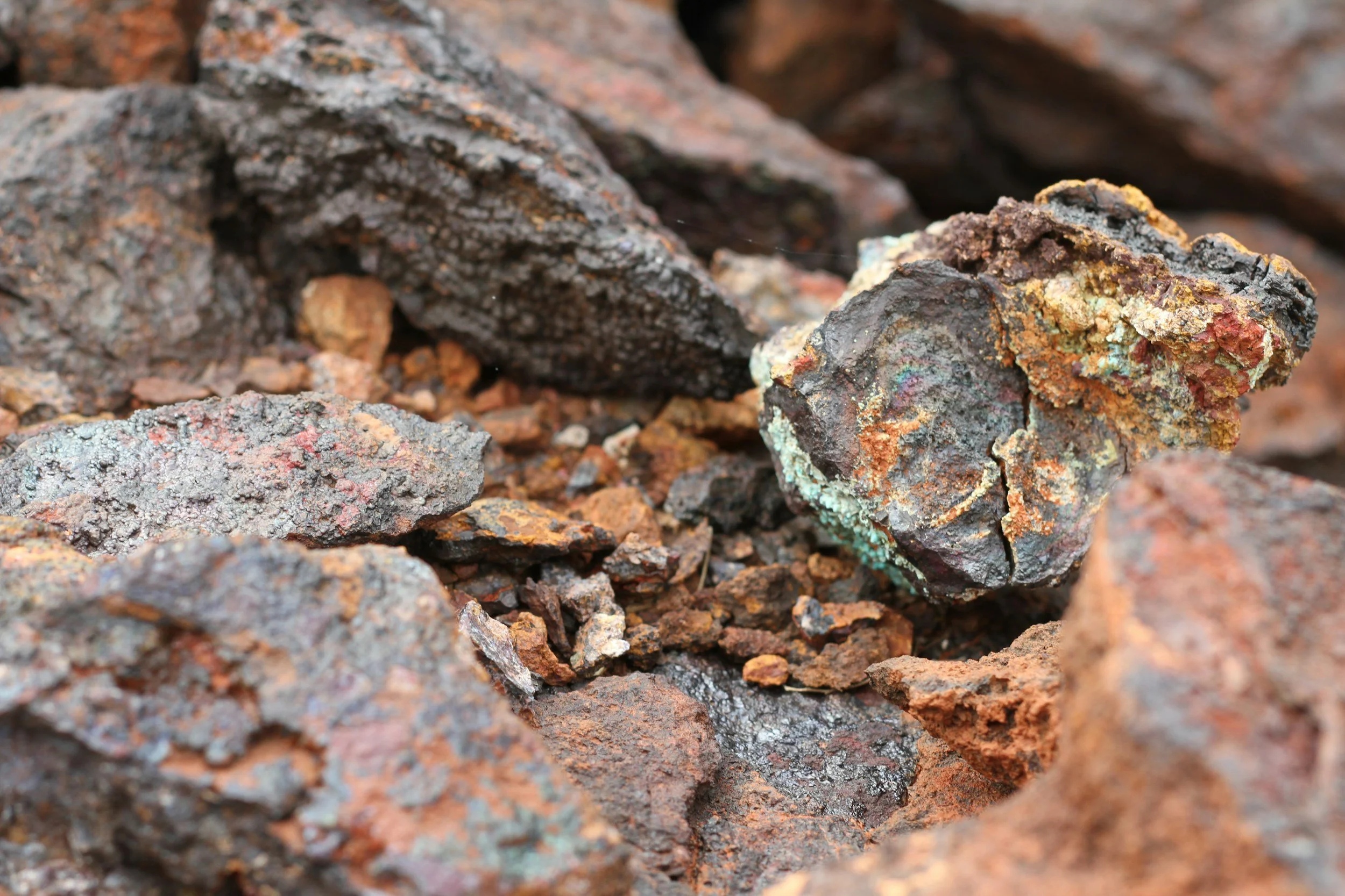 Close-up of a pile of rocks with various textures and colors, including dark gray, reddish-brown, and some patches of greenish-blue lichen.
