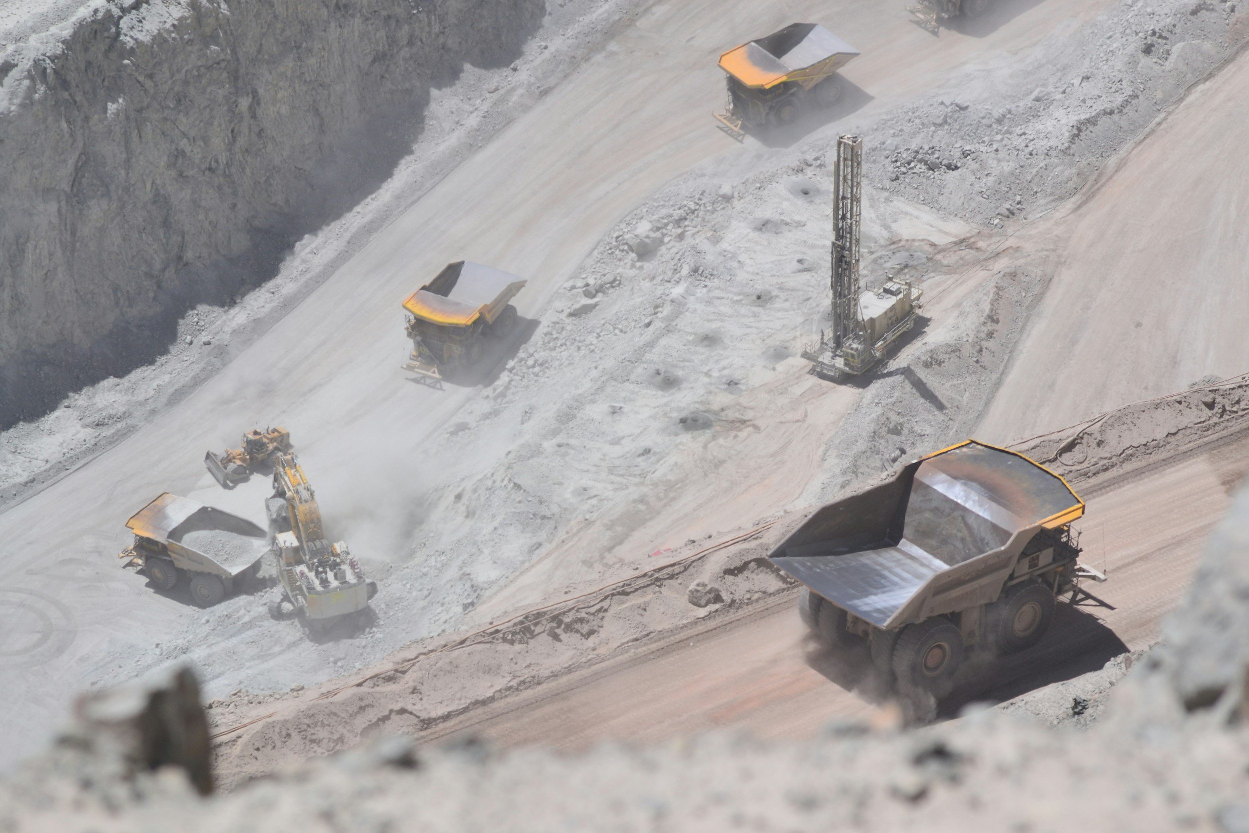 Heavy machinery working in a quarry, with dump trucks and a drilling rig on a dusty, excavated area.